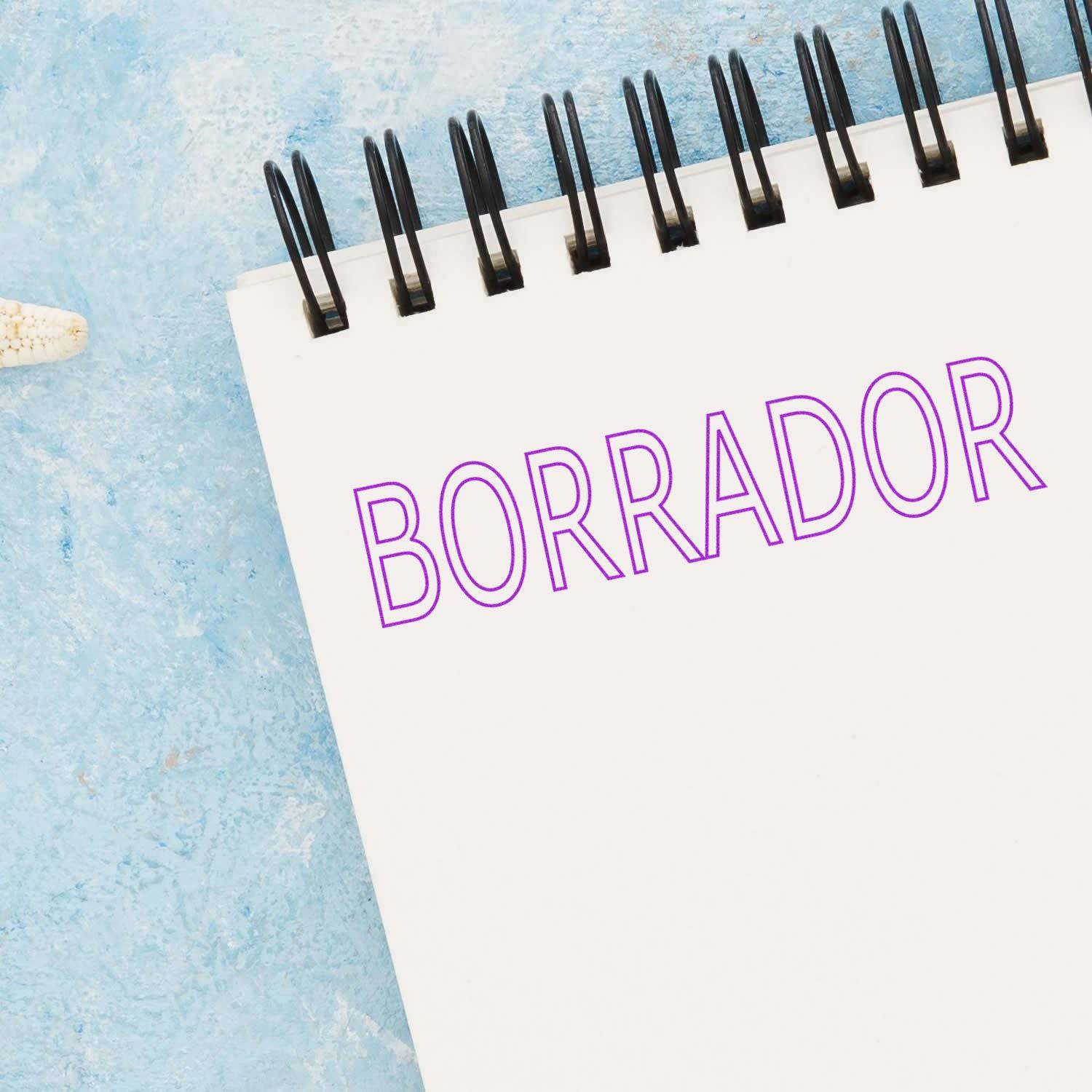 Large Pre-Inked Borrador Stamp in use on a white spiral notebook, displaying the word BORRADOR in purple ink against a blue background.