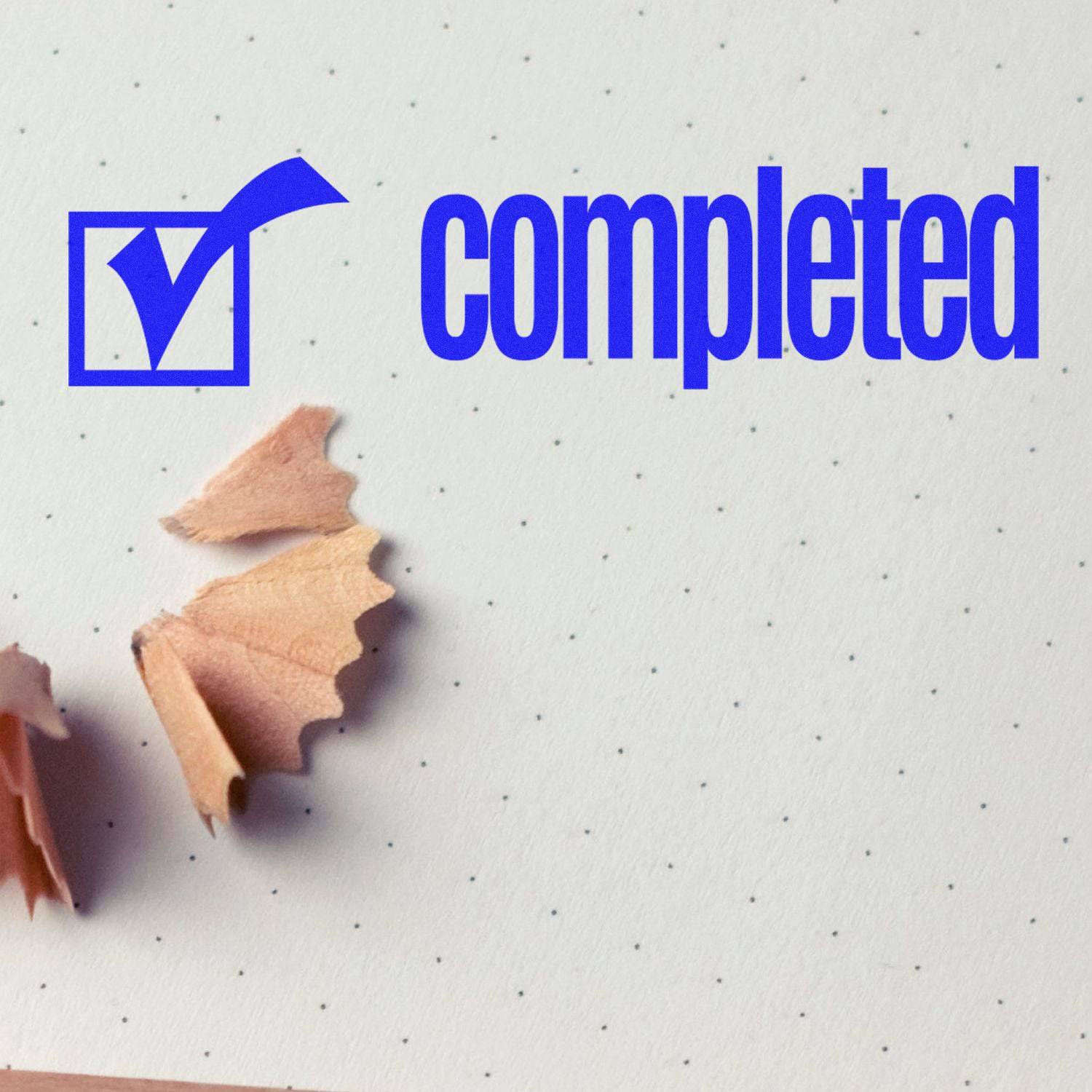 Large Self Inking Completed with Checkbox Stamp marking a dotted paper with a blue checkmark and 'completed' text, next to pencil shavings.