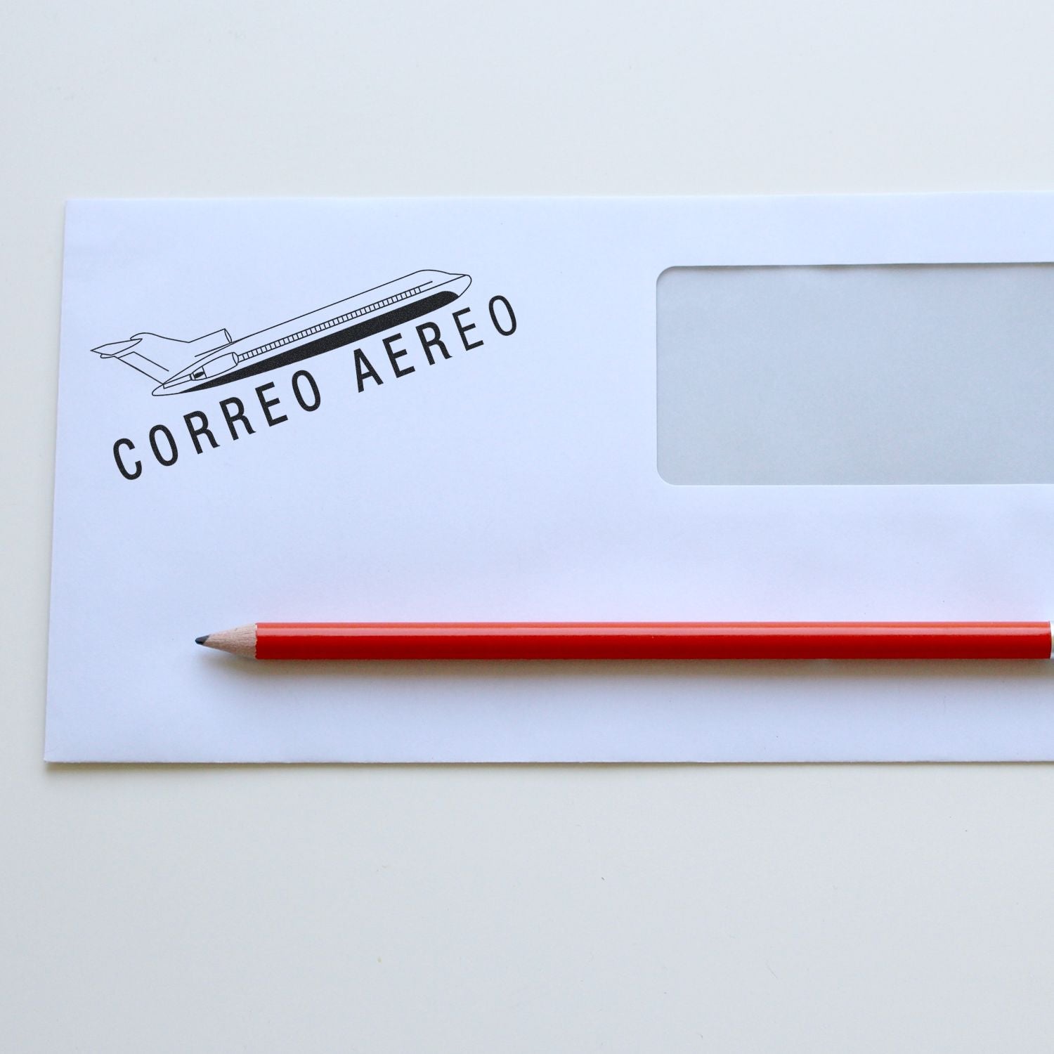 White envelope stamped with Large Correo Aero Rubber Stamp featuring an airplane design, accompanied by a red pencil on a white surface.