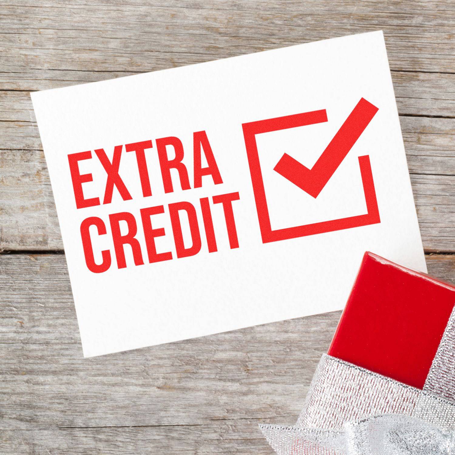 Large Pre-Inked Extra Credit Stamp marking a paper with a red Extra Credit checkmark, placed on a wooden surface next to a wrapped gift.