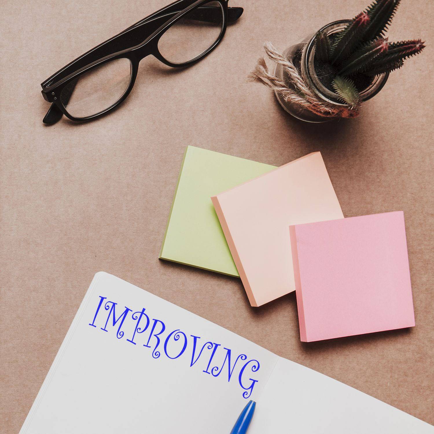 Large Pre-Inked Improving Stamp in use on a notebook, surrounded by colorful sticky notes, glasses, and a small potted plant.
