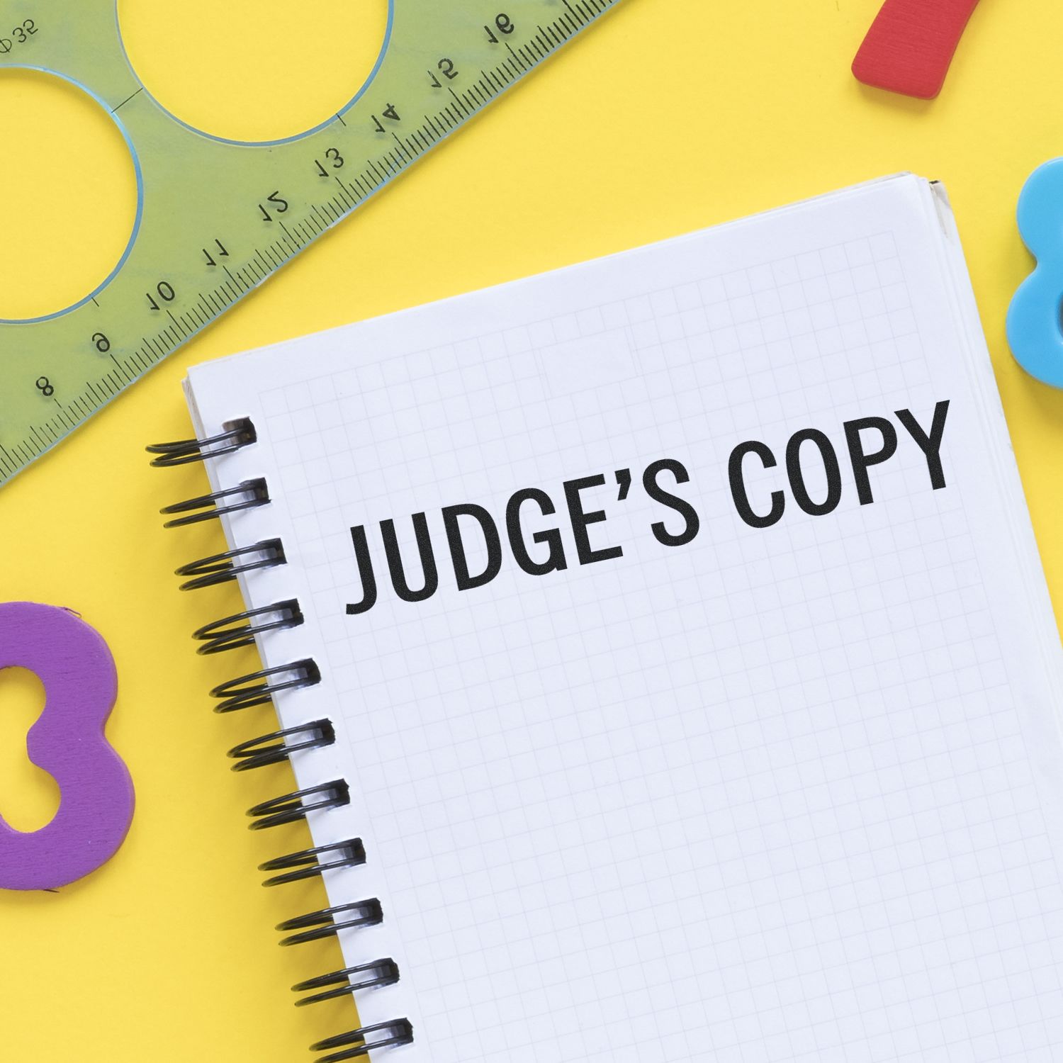 Self Inking Judge's Copy Stamp used on a spiral notebook with JUDGE'S COPY text, surrounded by colorful stationery on a yellow background.