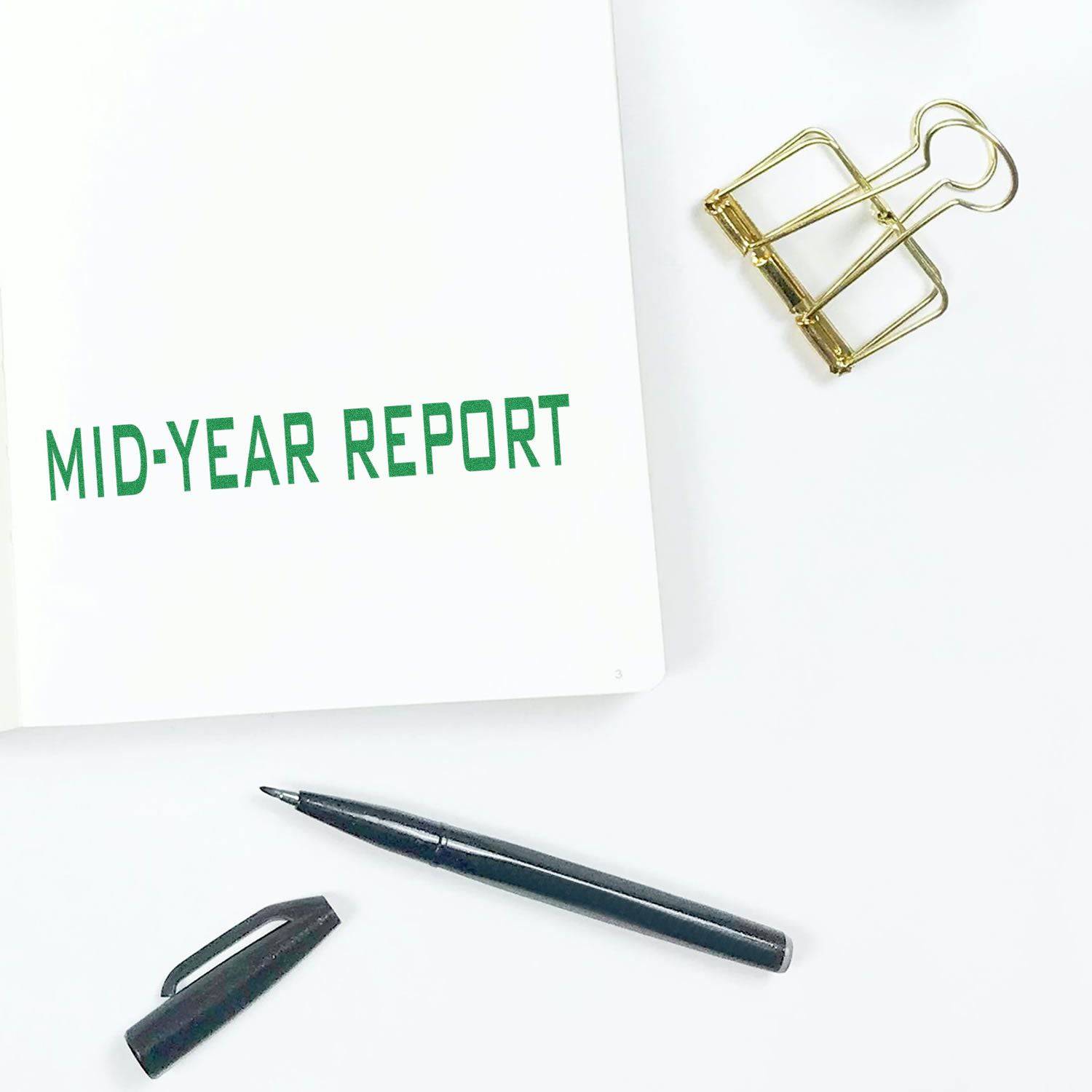 Large Pre-Inked Mid-Year Report Stamp used on a white paper, with a black pen, pen cap, and gold binder clip nearby.