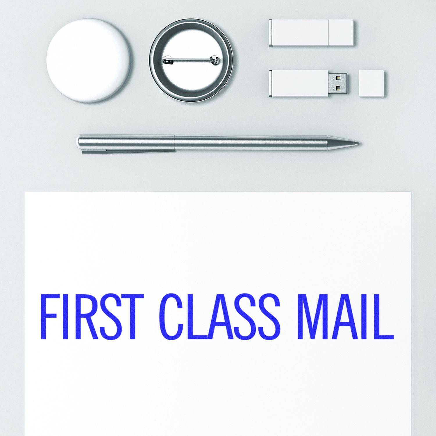 Large Pre-Inked Narrow First Class Mail Stamp used on white paper, surrounded by a pen, USB drives, and round objects on a white surface.