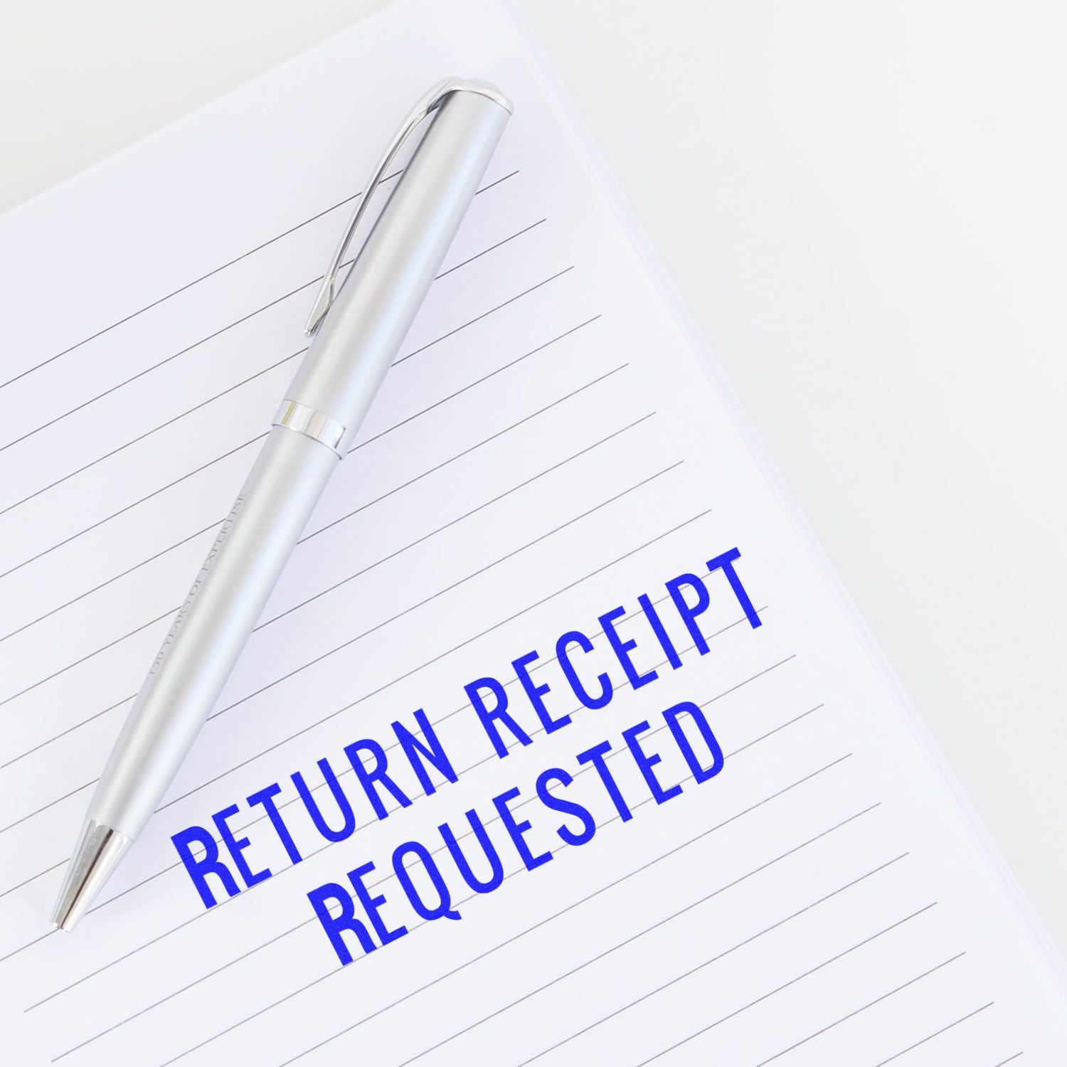 Narrow Font Return Receipt Requested Rubber Stamp in blue ink on lined paper with a silver pen placed beside it.
