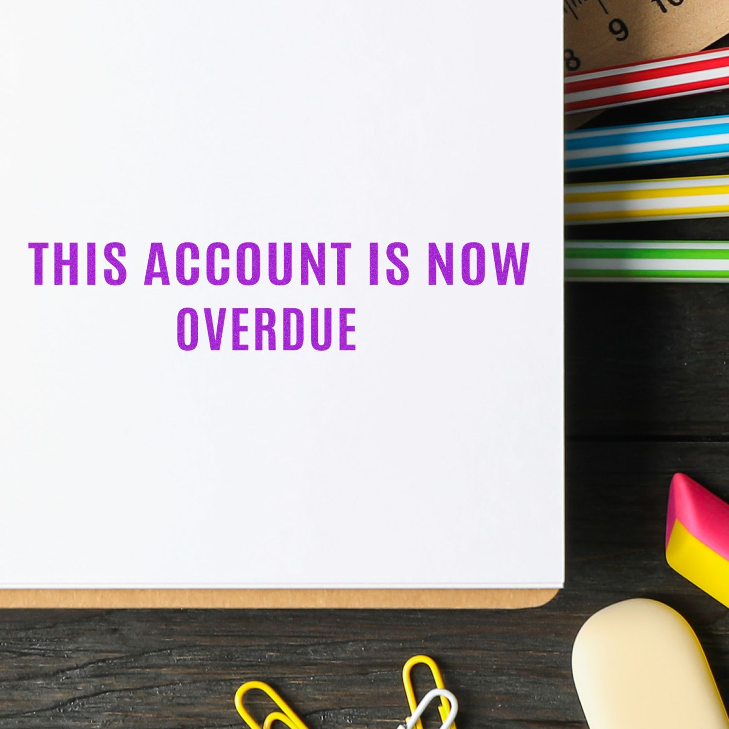 Large Narrow This Account Is Now Overdue Rubber Stamp in use on a white paper, surrounded by colorful office supplies on a dark wooden desk.
