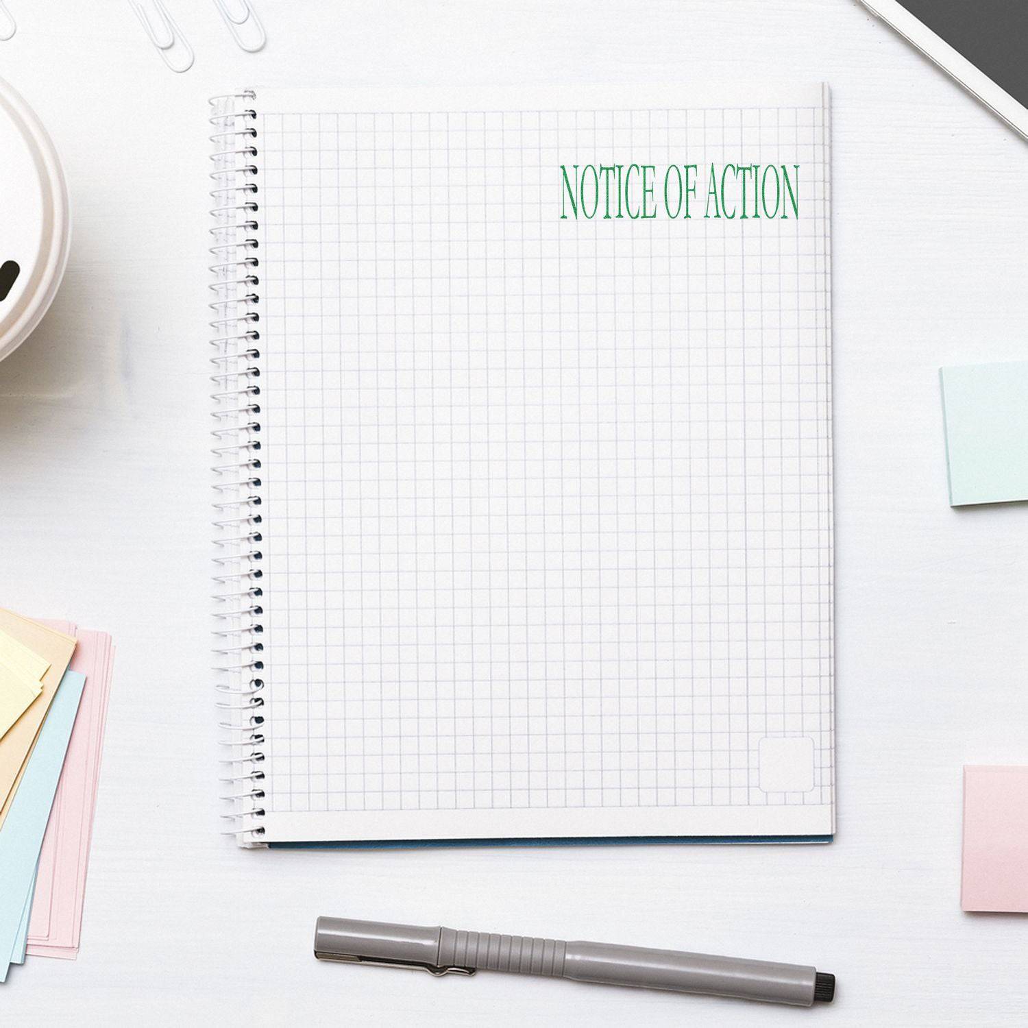 Large Pre-Inked Notice Of Action Stamp used on a spiral notebook with grid paper, surrounded by a pen, sticky notes, and a smartphone.