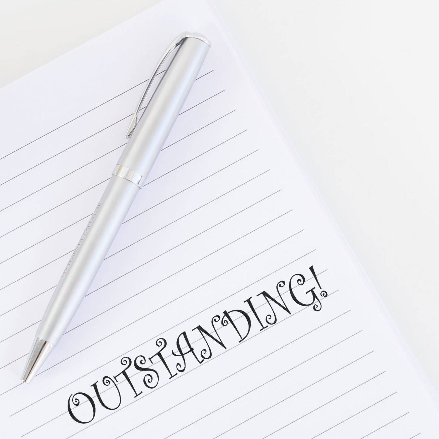 A silver pen rests on a lined notebook with the word OUTSTANDING! stamped in bold using the Large Pre-Inked Outstanding Stamp.