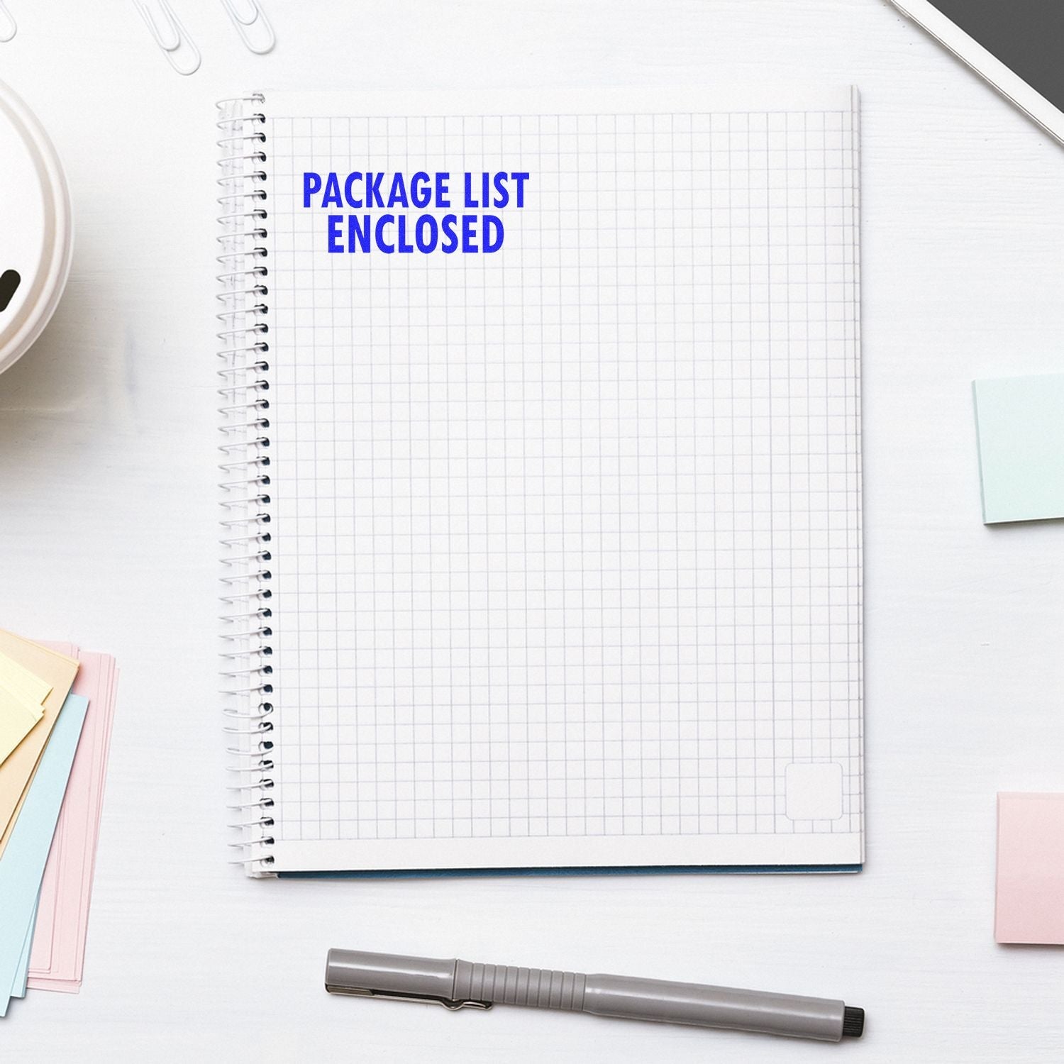 Self Inking Package List Enclosed Shipping Stamp used on a grid notebook, surrounded by office supplies on a white desk.