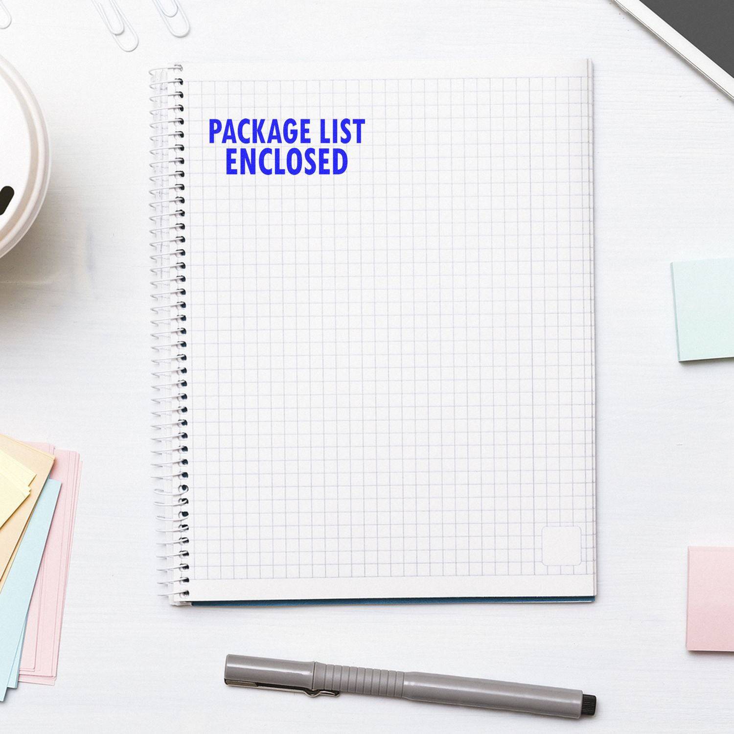 Large Package List Enclosed Rubber Stamp used on a spiral notebook with grid paper, surrounded by a pen, sticky notes, and a phone.