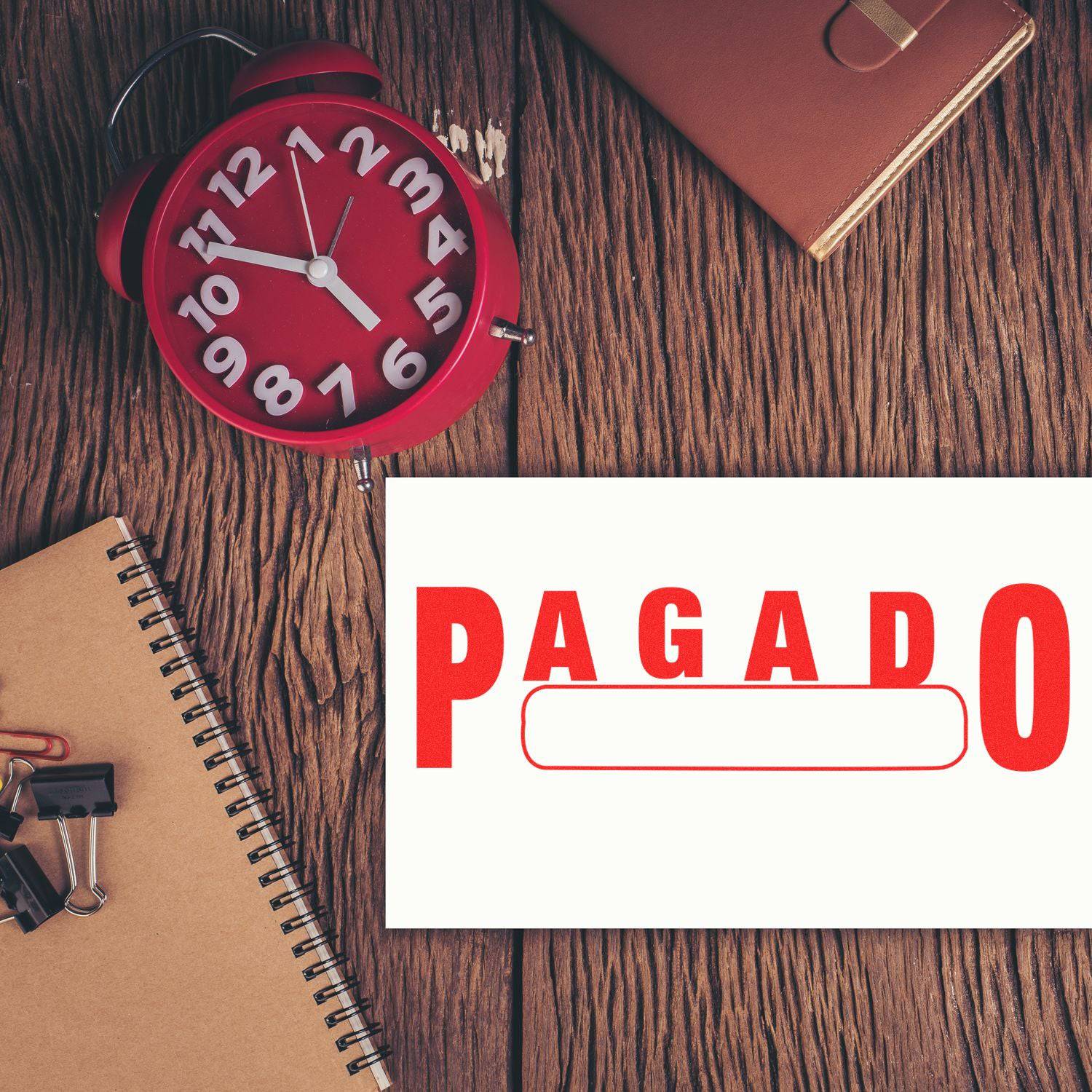 Large Pre-Inked Pagado with Box Stamp on a wooden desk with a red clock, notebook, and office supplies.