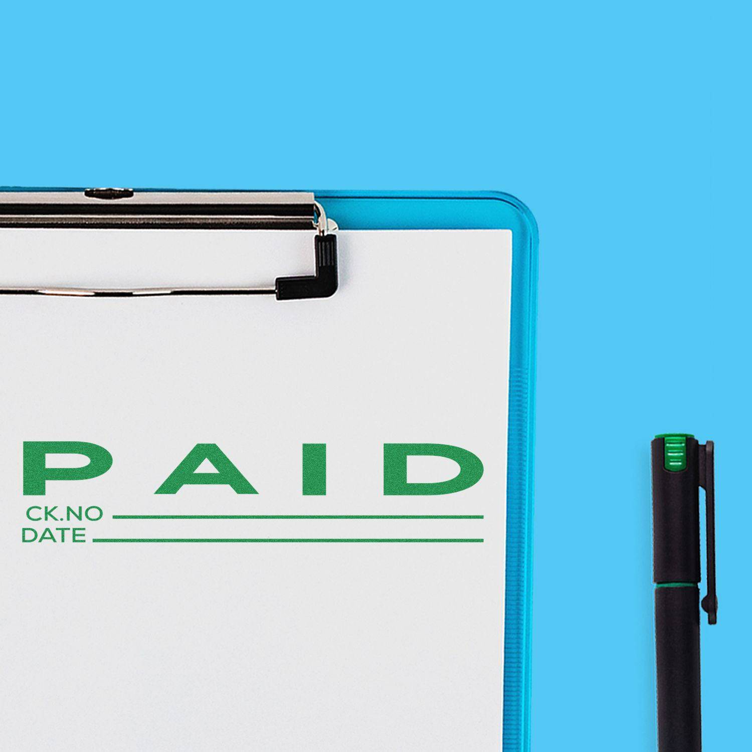 Large Pre-Inked Paid with Ck No Stamp in green ink on a white paper clipped to a blue clipboard, with a black pen beside it.