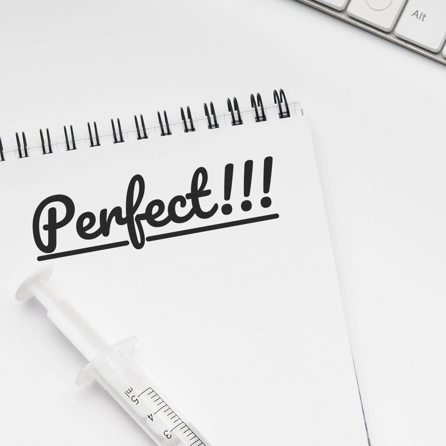 Large Perfect Rubber Stamp imprint reading Perfect!!! on a spiral notebook, with a syringe pen and keyboard in the background.