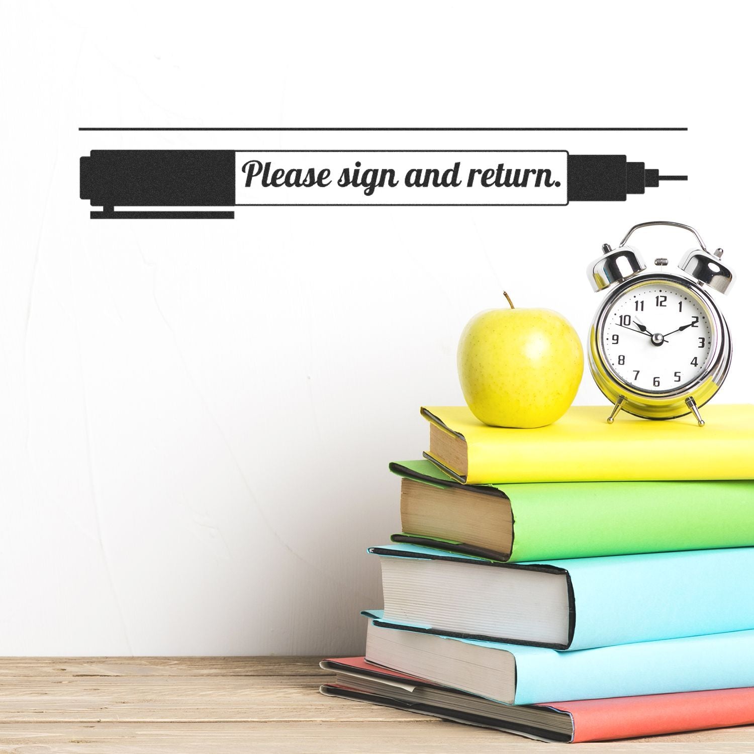 Slim Pre-Inked Please Sign and Return with Pen Stamp on a white wall above a stack of colorful books, an apple, and an alarm clock.
