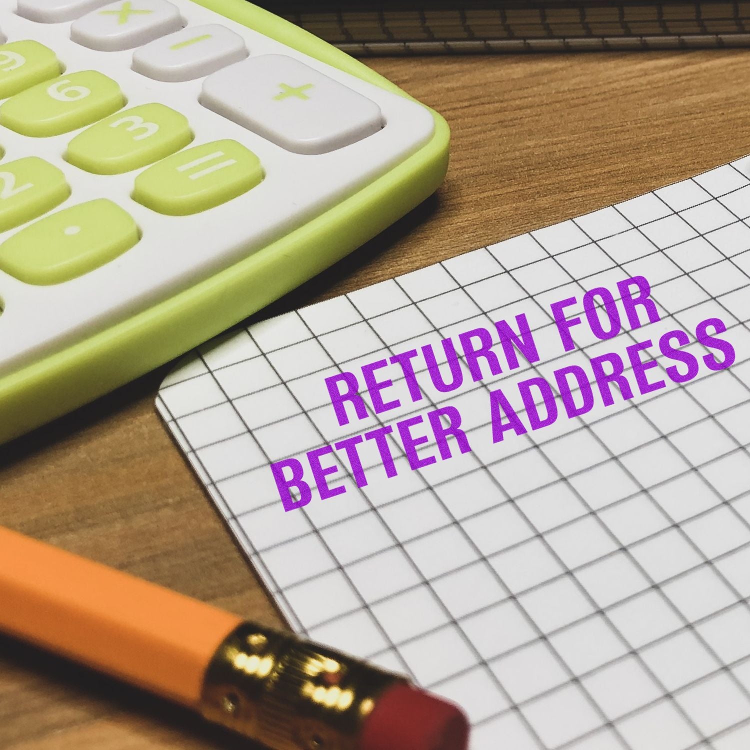 Large Self Inking Return for Better Address Stamp used on a grid paper, next to a calculator and pencil on a wooden desk.