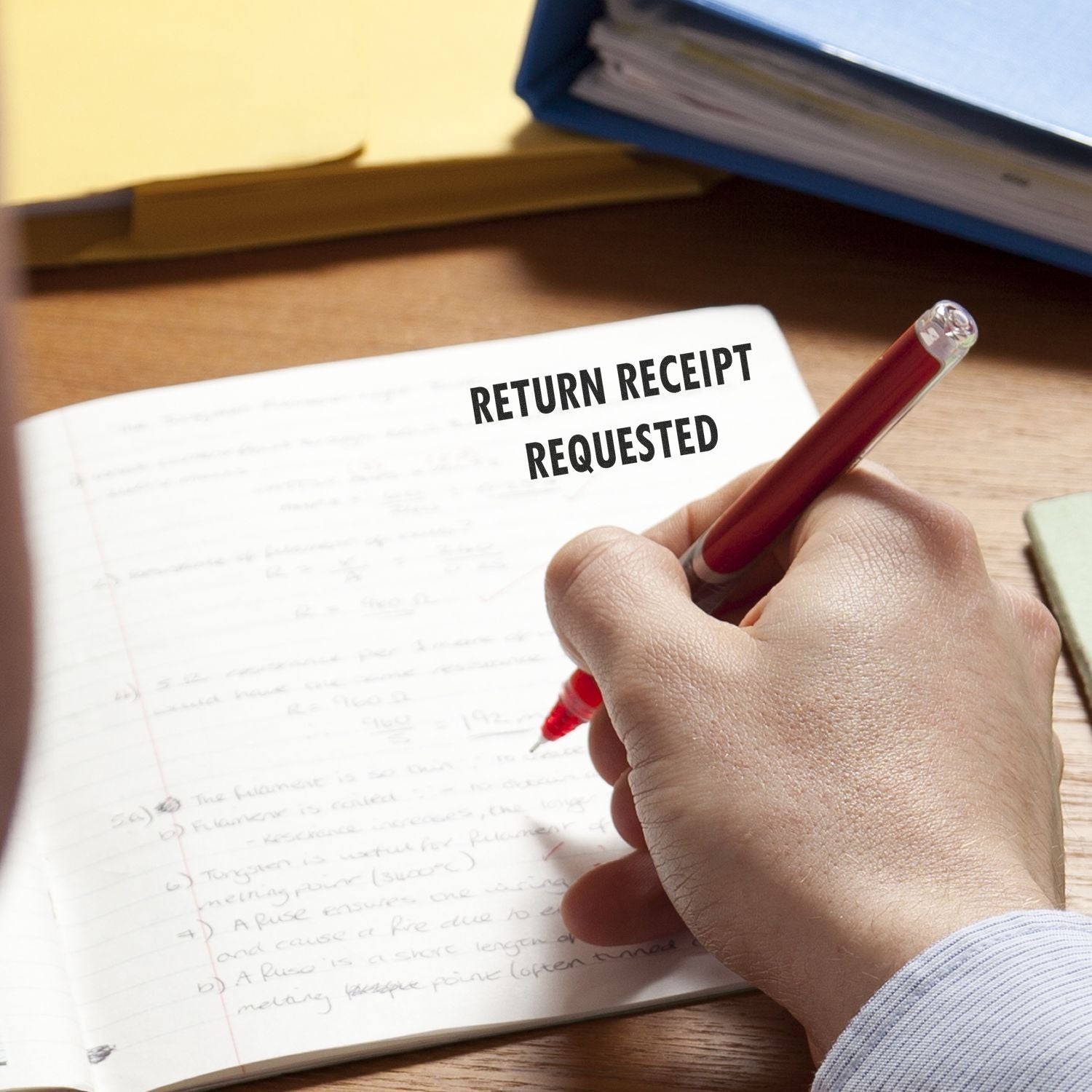 A hand writing in a notebook with a red pen, next to a Return Receipt Requested rubber stamp impression on the page.