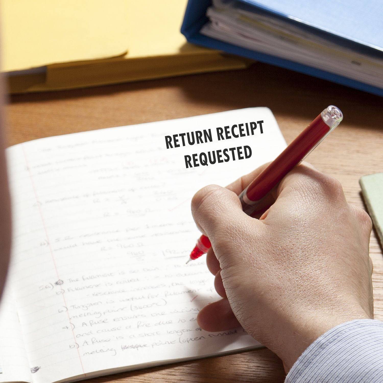 A hand using the Large Pre-Inked Return Receipt Requested Stamp on a notebook, with folders and papers in the background.