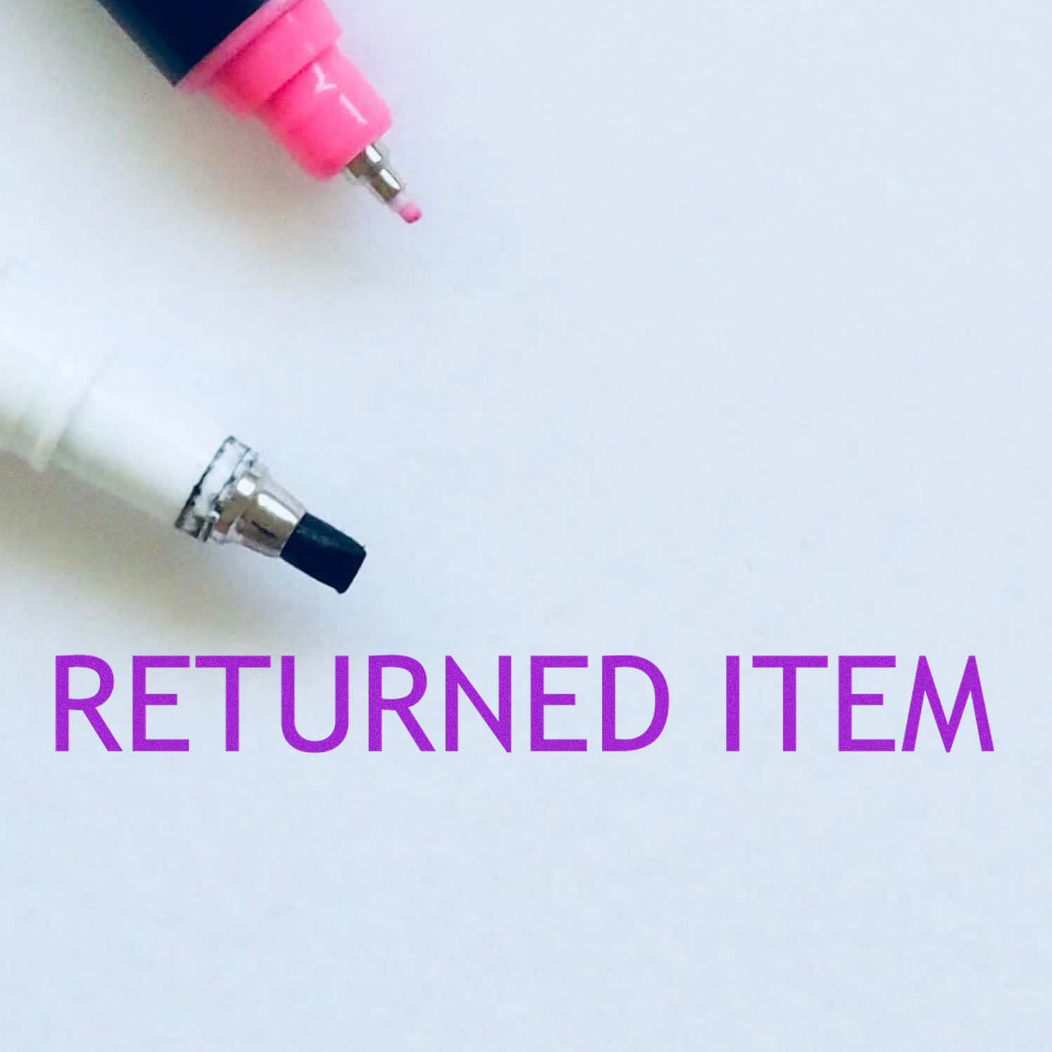 Slim Pre-Inked Returned Item Stamp in use, marking RETURNED ITEM in purple on white paper, with two pens nearby.