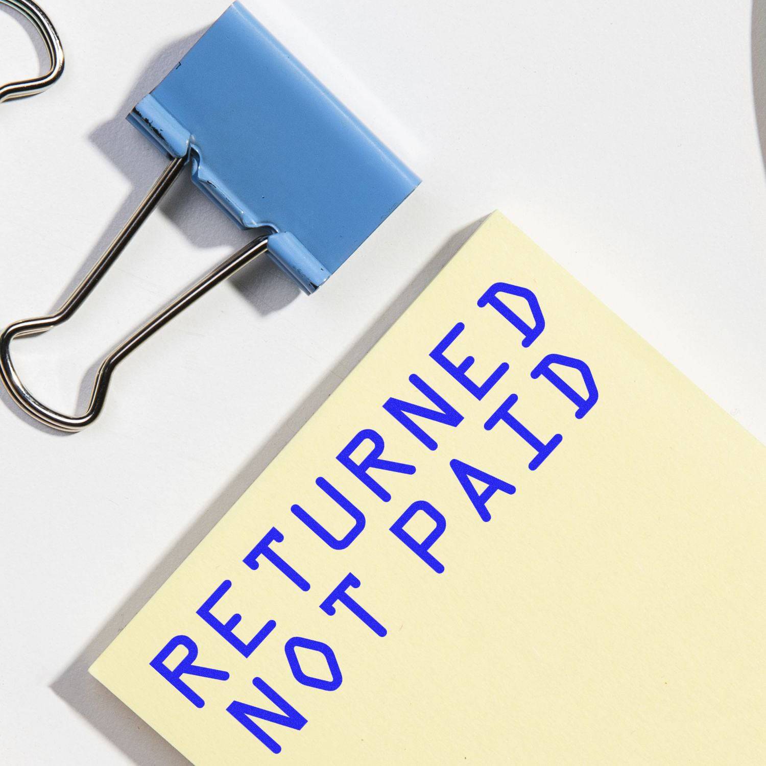 A blue Large Pre-Inked Returned Not Paid Stamp on a yellow paper next to a blue binder clip on a white surface.