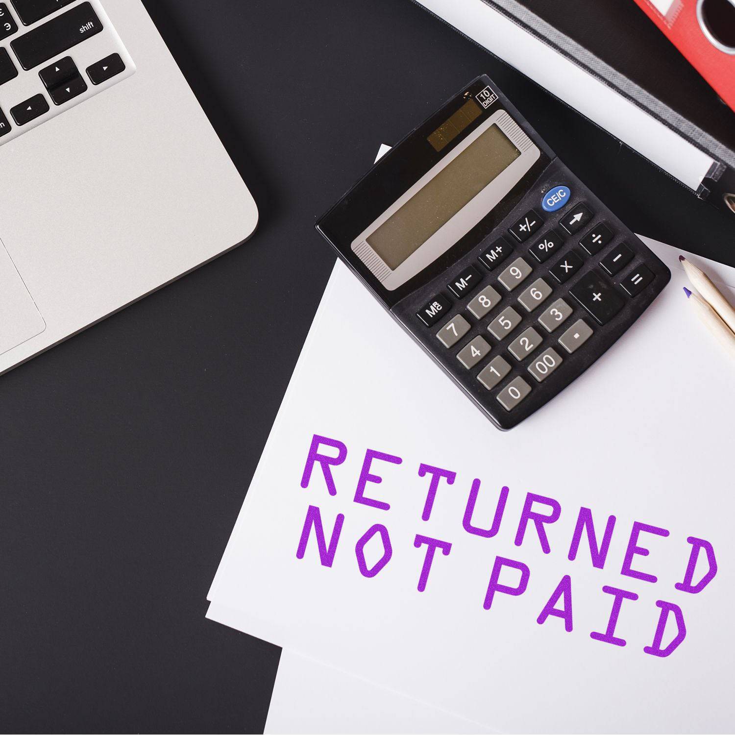 Large Pre-Inked Returned Not Paid Stamp used on white paper next to a calculator, laptop, and pencil on a black desk.