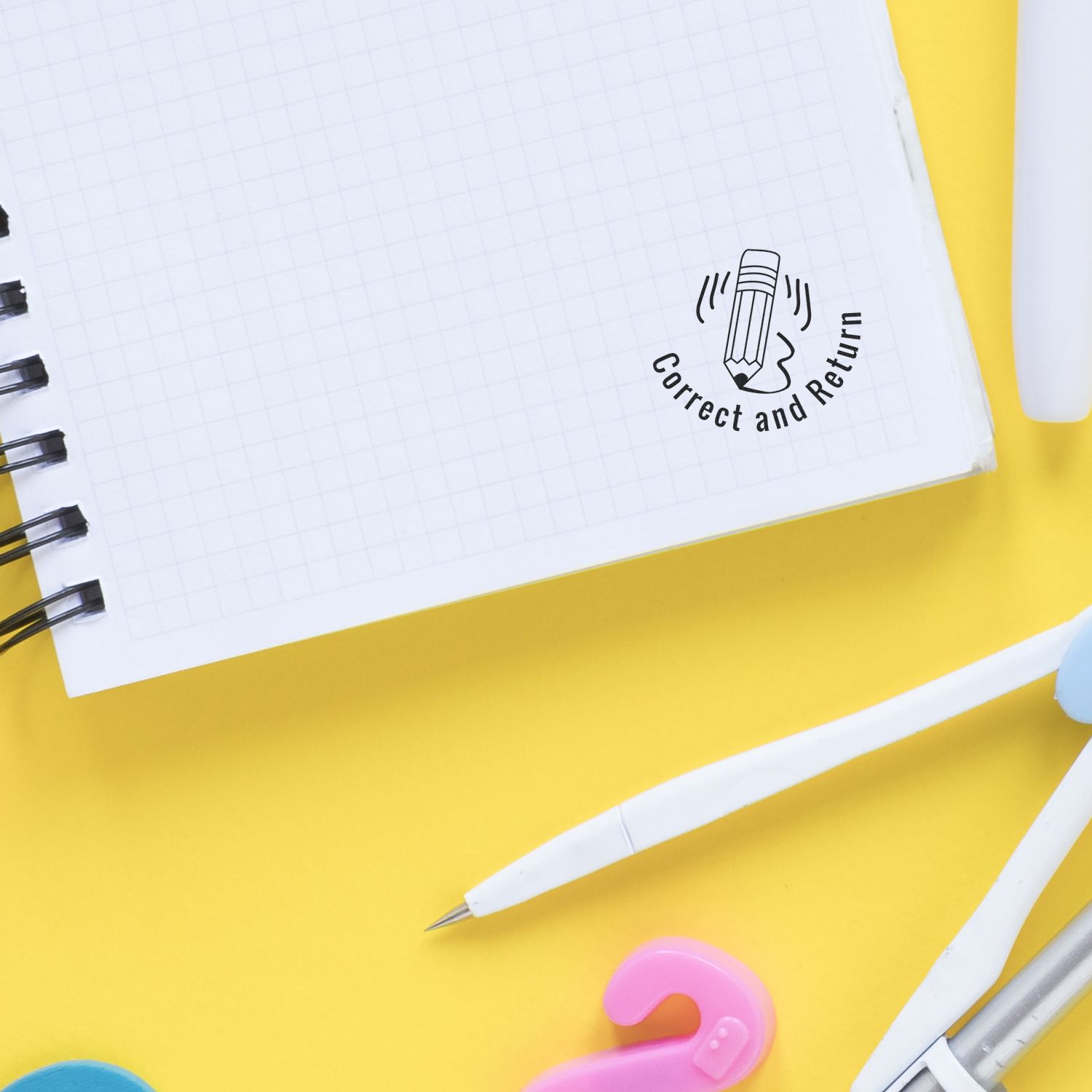 Self Inking Round Correct and Return Stamp on a notebook page with a pen, eraser, and paper clips on a yellow background.