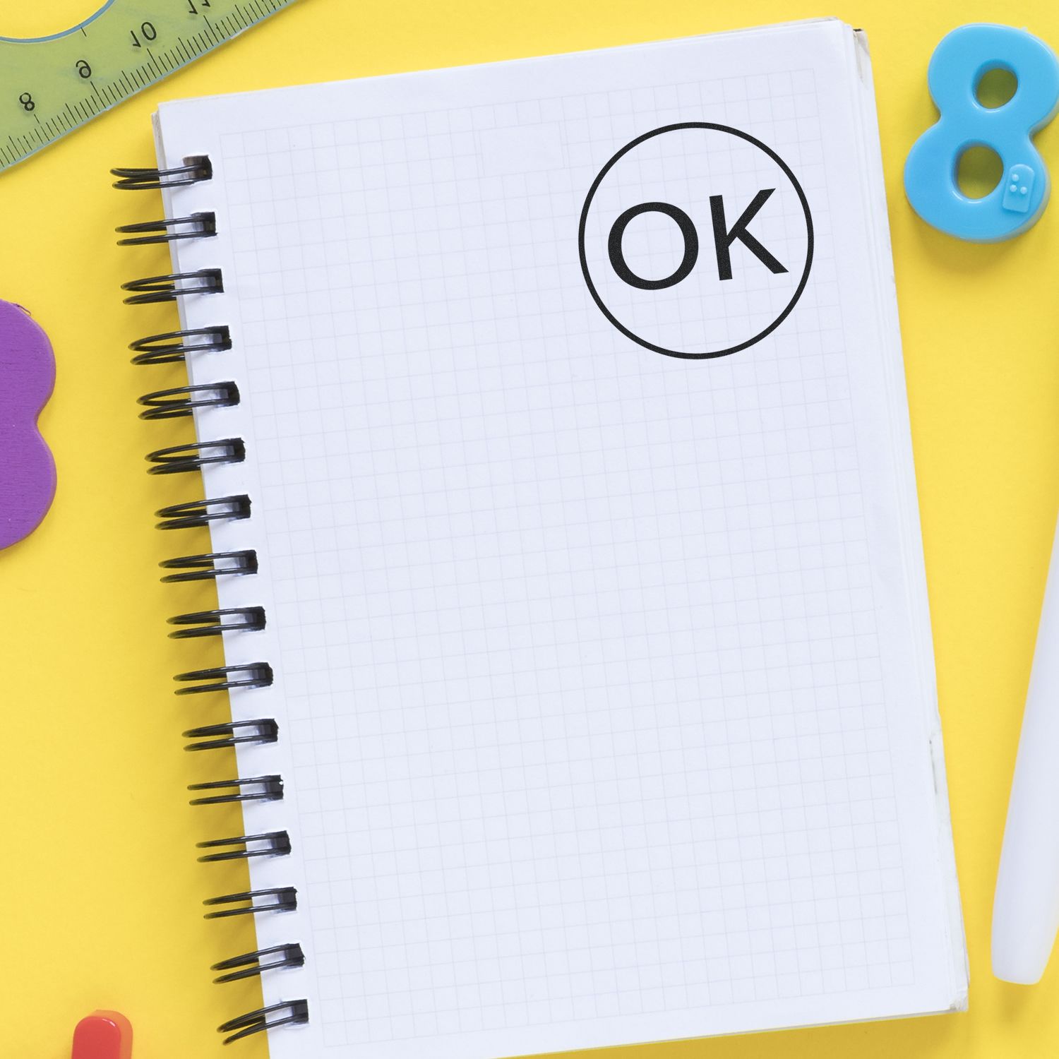 Self Inking Round OK Stamp marking a grid notebook page, surrounded by colorful stationery on a yellow background.