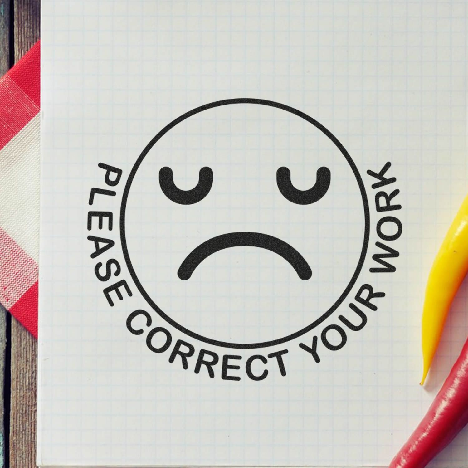 Self Inking Round Please Correct Smiley Stamp on graph paper with a sad face and Please Correct Your Work text, next to a red and yellow pepper.
