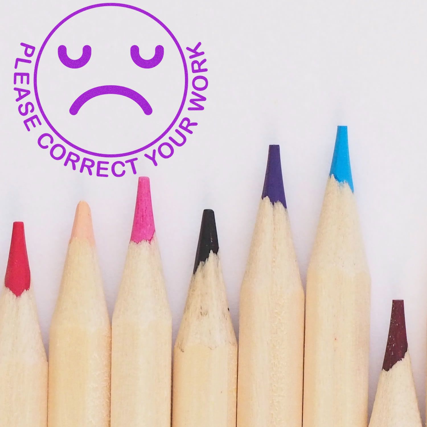 Round Please Correct Your Work smiley rubber stamp in purple ink next to a row of sharpened colored pencils.