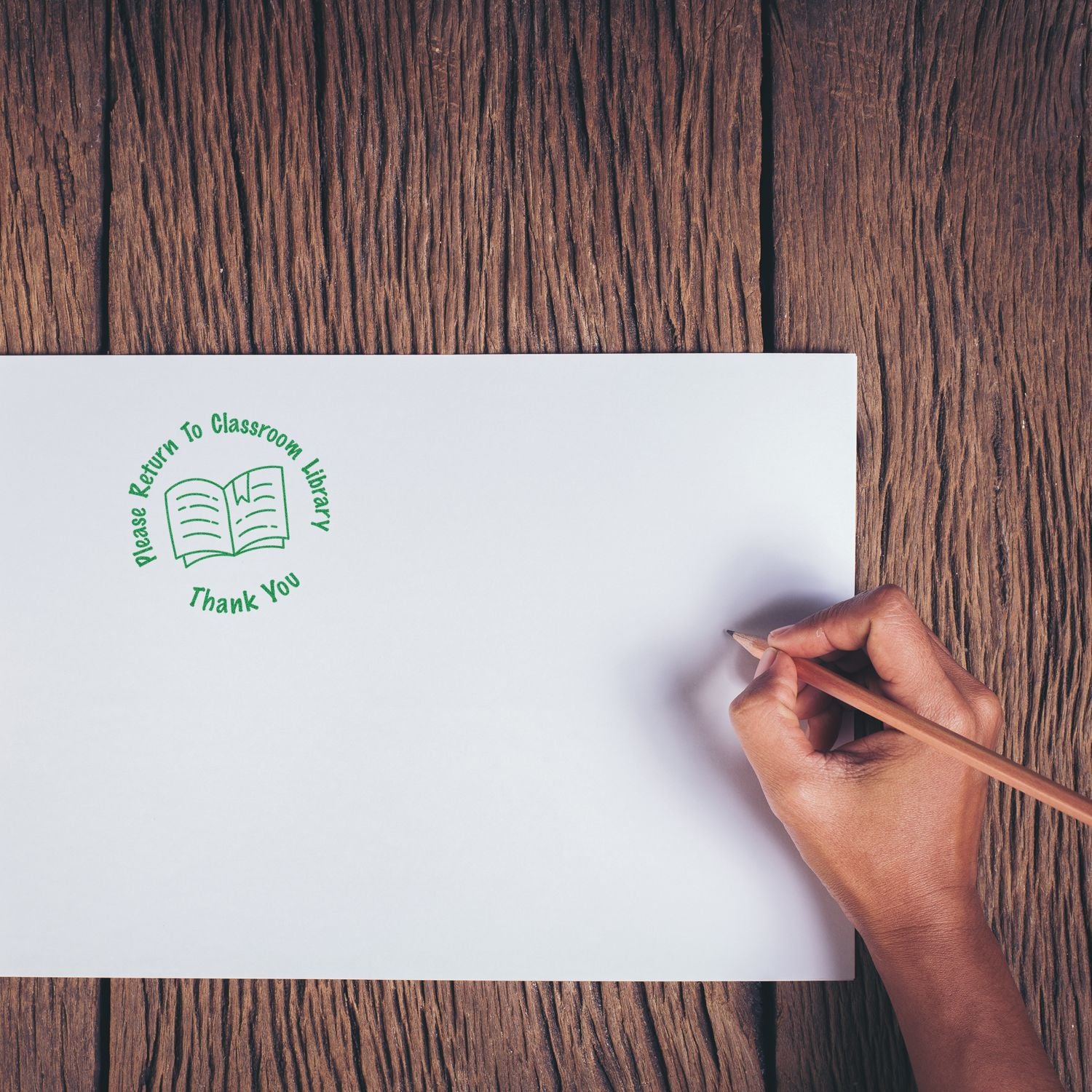 Hand holding pencil next to a green Round Please Return to Classroom Library Rubber Stamp imprint on a white paper on wooden table.