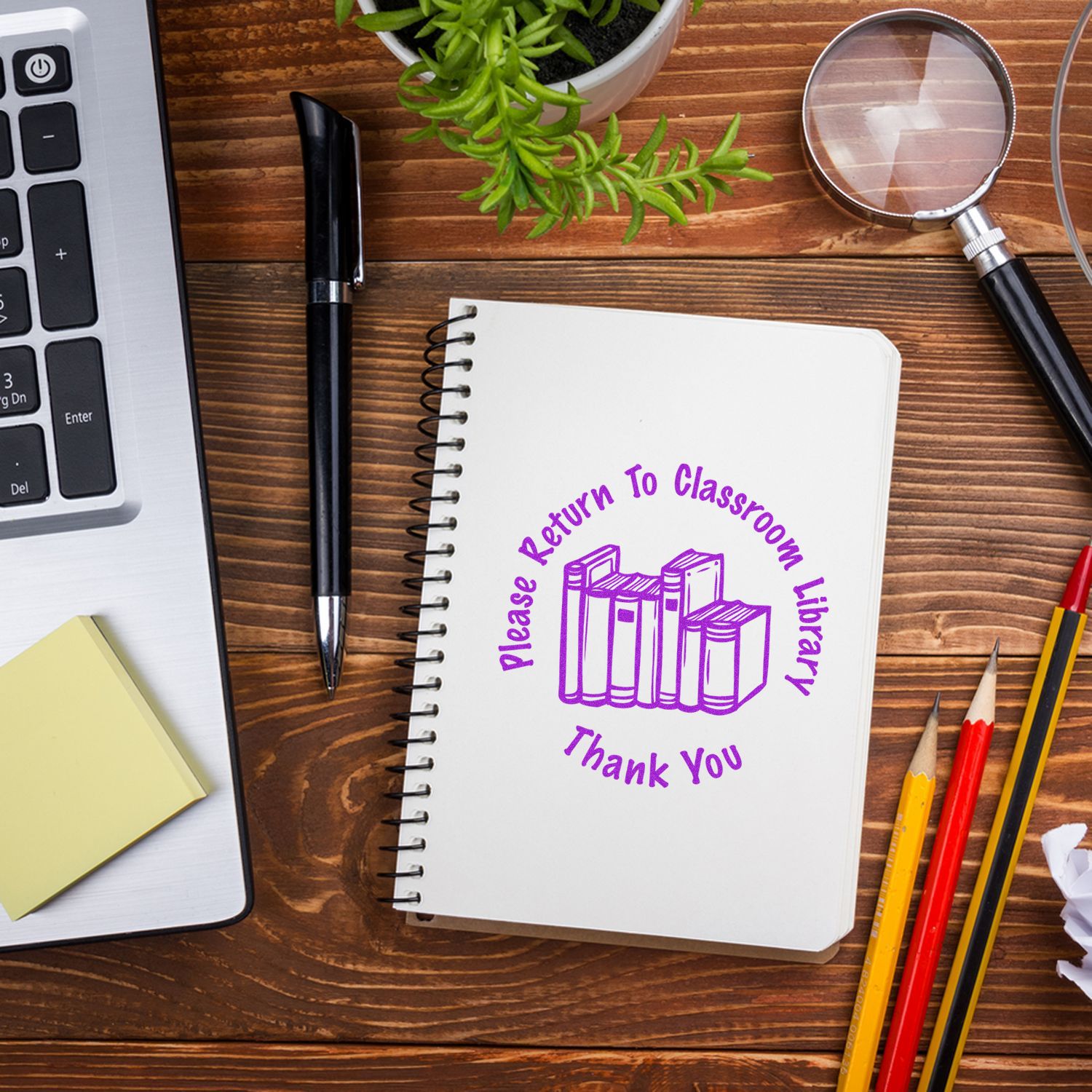 Notebook stamped with Self Inking Round Please Return to Classroom Stamp on a wooden desk with a laptop, pen, magnifying glass, and pencils.