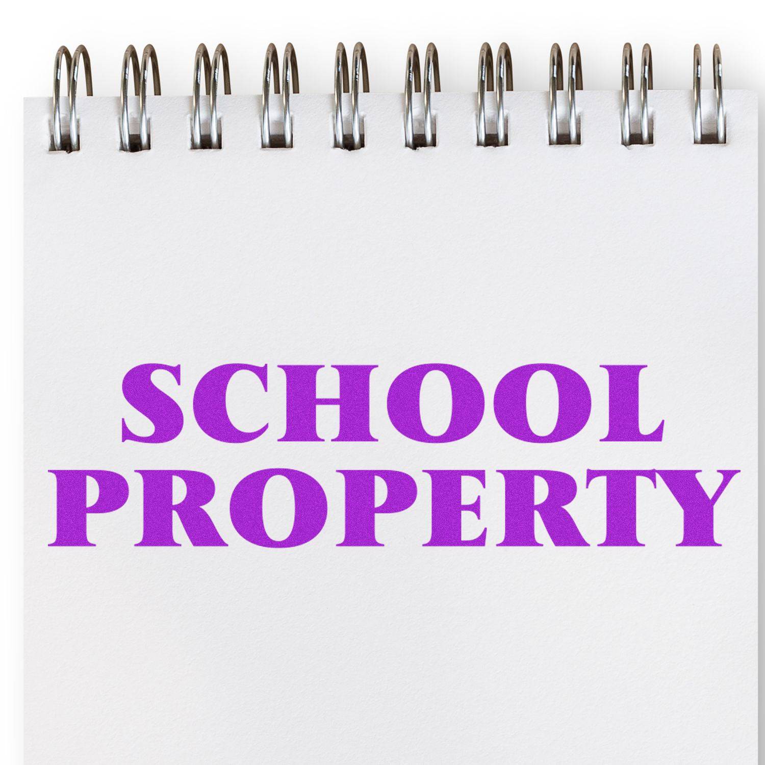 Large School Property Rubber Stamp in use, stamping SCHOOL PROPERTY in bold purple letters on a white spiral-bound notebook page.