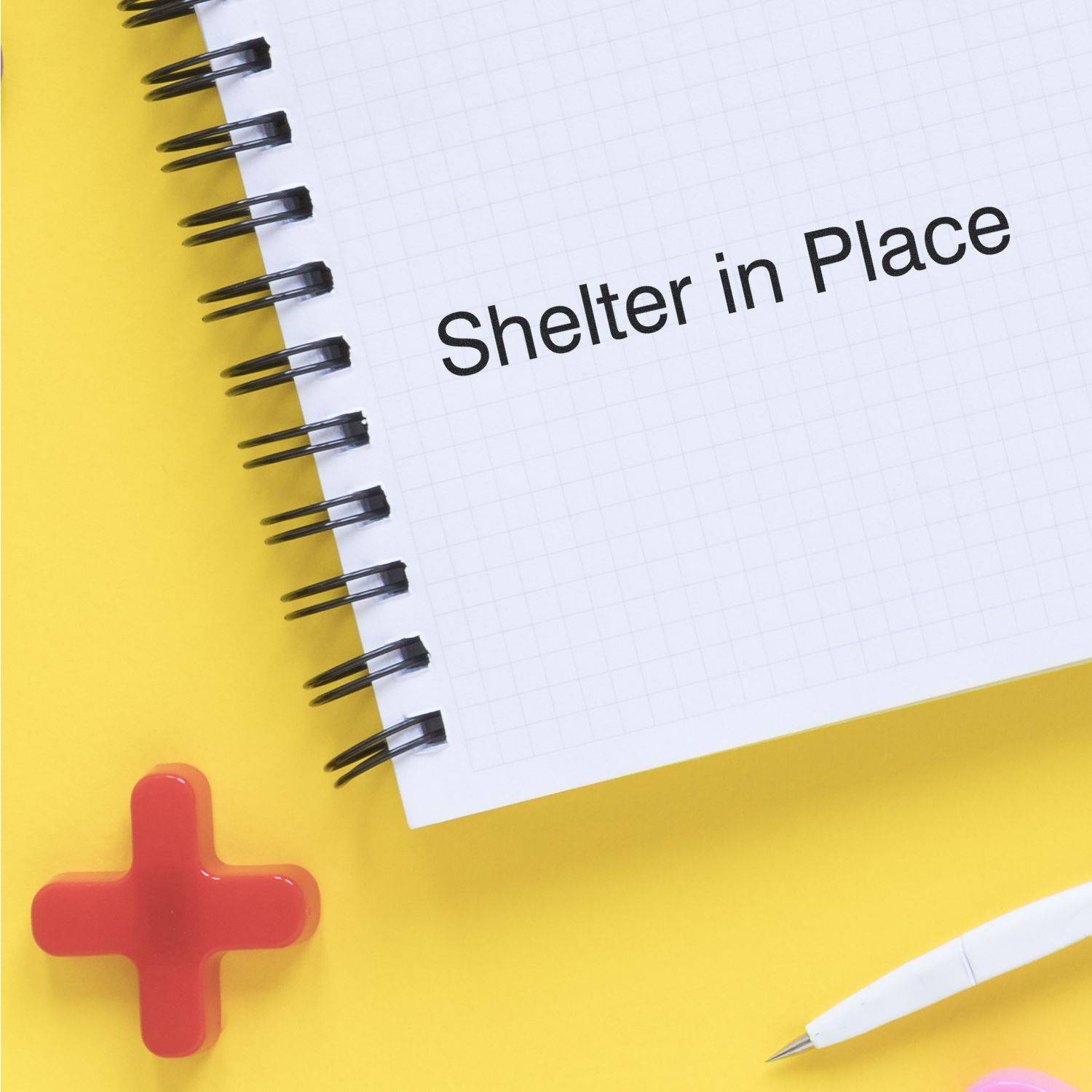 Large Pre-Inked Shelter in Place Stamp used on a spiral notebook with Shelter in Place text, placed on a yellow surface with a pen and red cross.