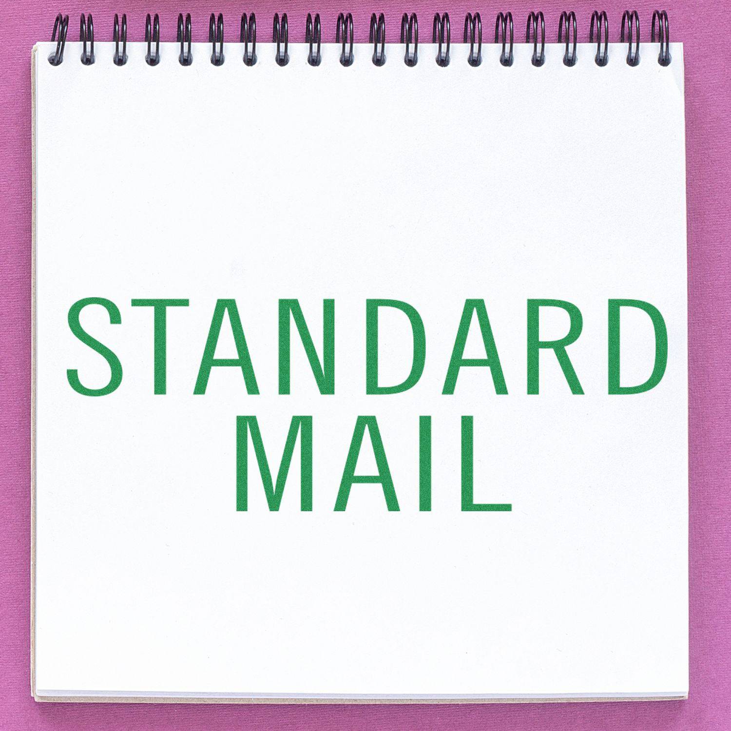 Large Pre-Inked Standard Mail Stacked Stamp imprint on a white notepad with green text, placed on a pink surface.