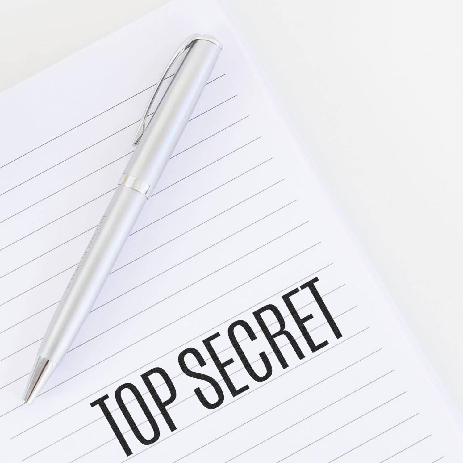 A silver pen rests on a lined notebook with TOP SECRET stamped in bold black letters using the Large Pre-Inked Top Secret Stamp.