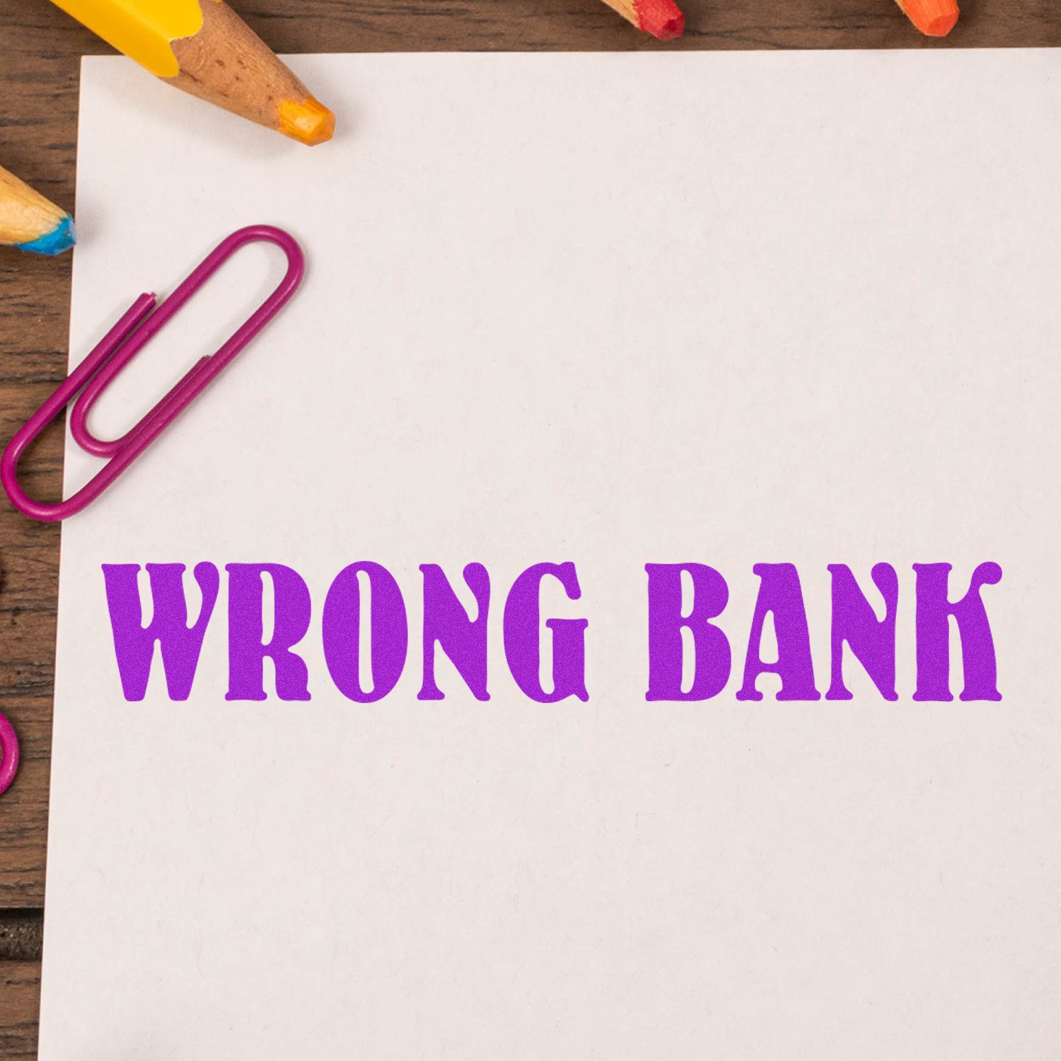 Purple 'Wrong Bank' rubber stamp on white paper, surrounded by colorful pencils and a pink paperclip on a wooden surface.