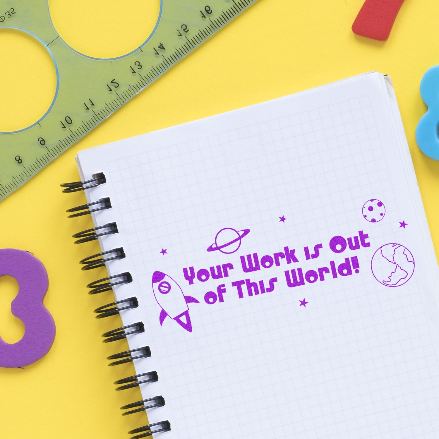 Spiral notebook stamped with Your Work is Out of This World in purple ink, surrounded by colorful stationery on a yellow background.