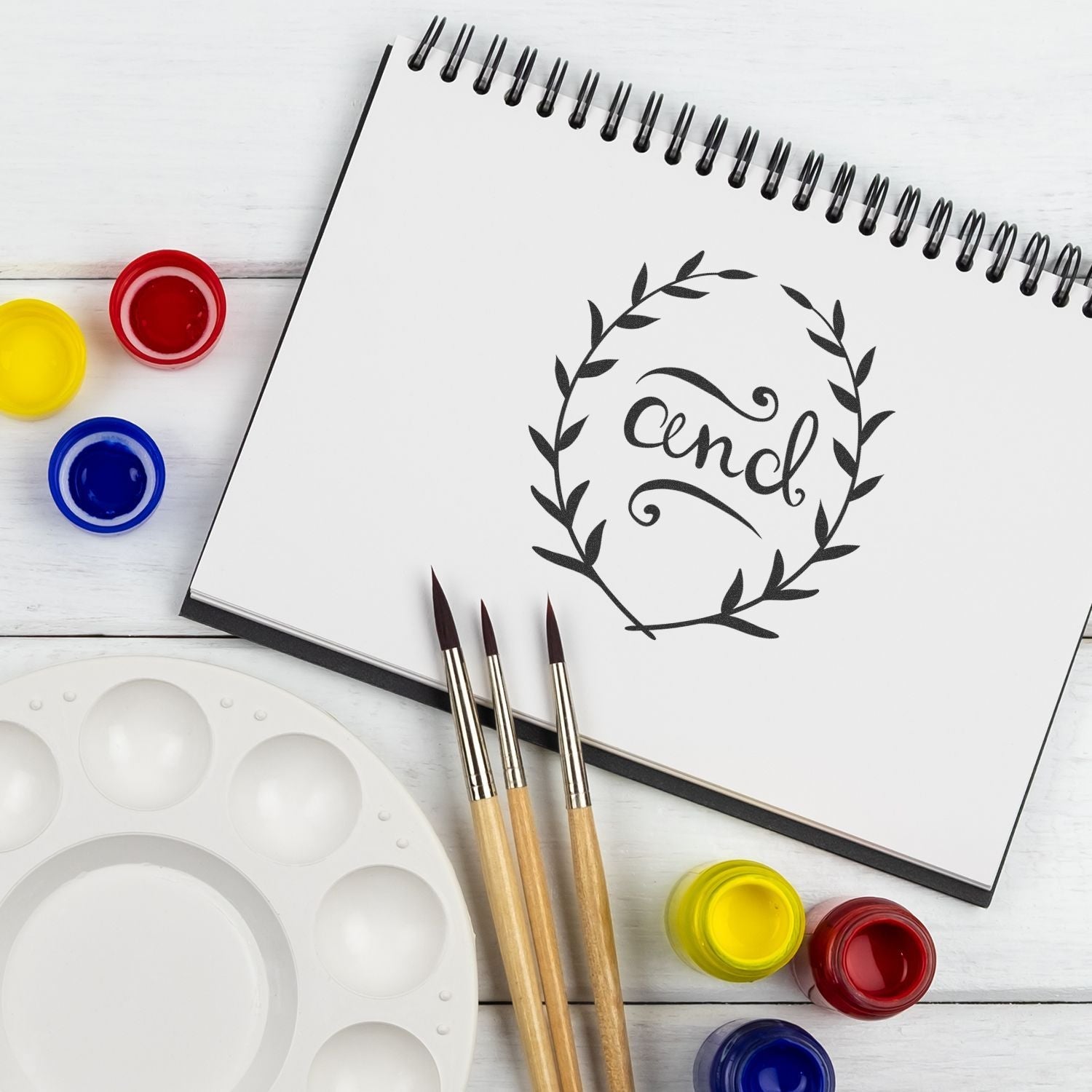 A Custom Rubber Stamp Size 4 Inch Diameter design on a sketchbook, surrounded by paintbrushes, paint pots, and a palette on a white surface.