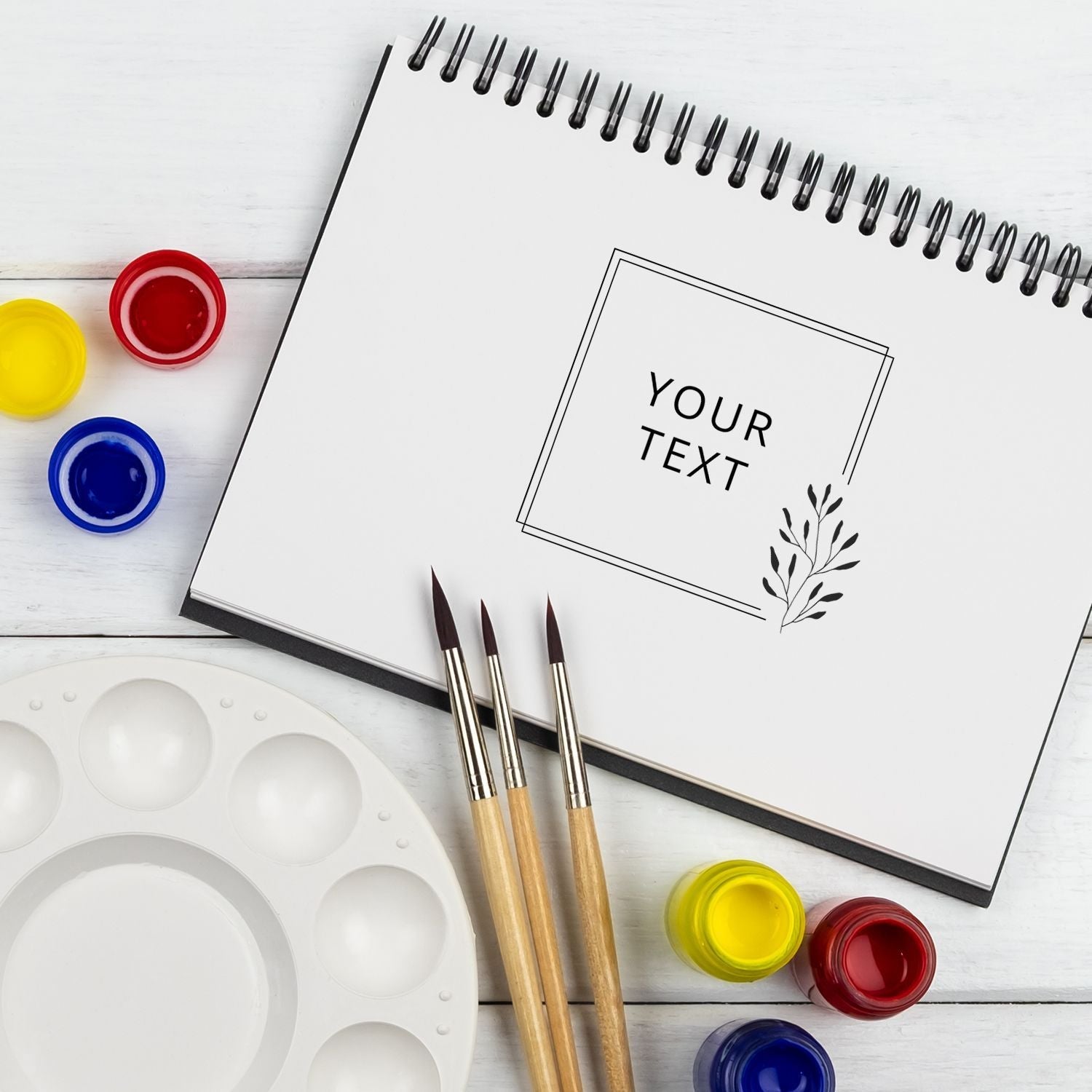 Custom Rubber Stamp Size 4 x 4 in use on a spiral notebook with paintbrushes, paint pots, and a palette on a white wooden surface.