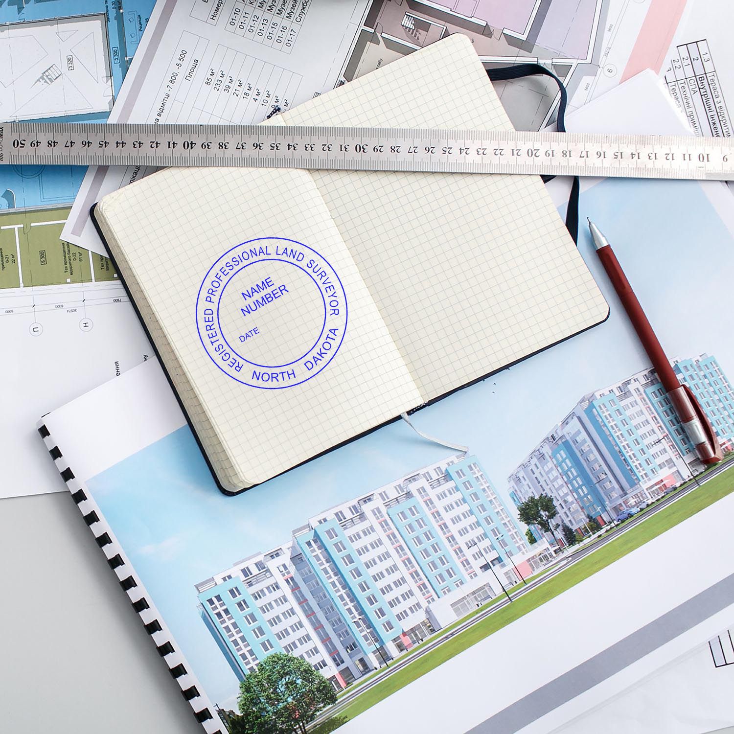 Self Inking North Dakota Land Surveyor Stamp in use on a notebook, surrounded by architectural plans, a ruler, and a pen.