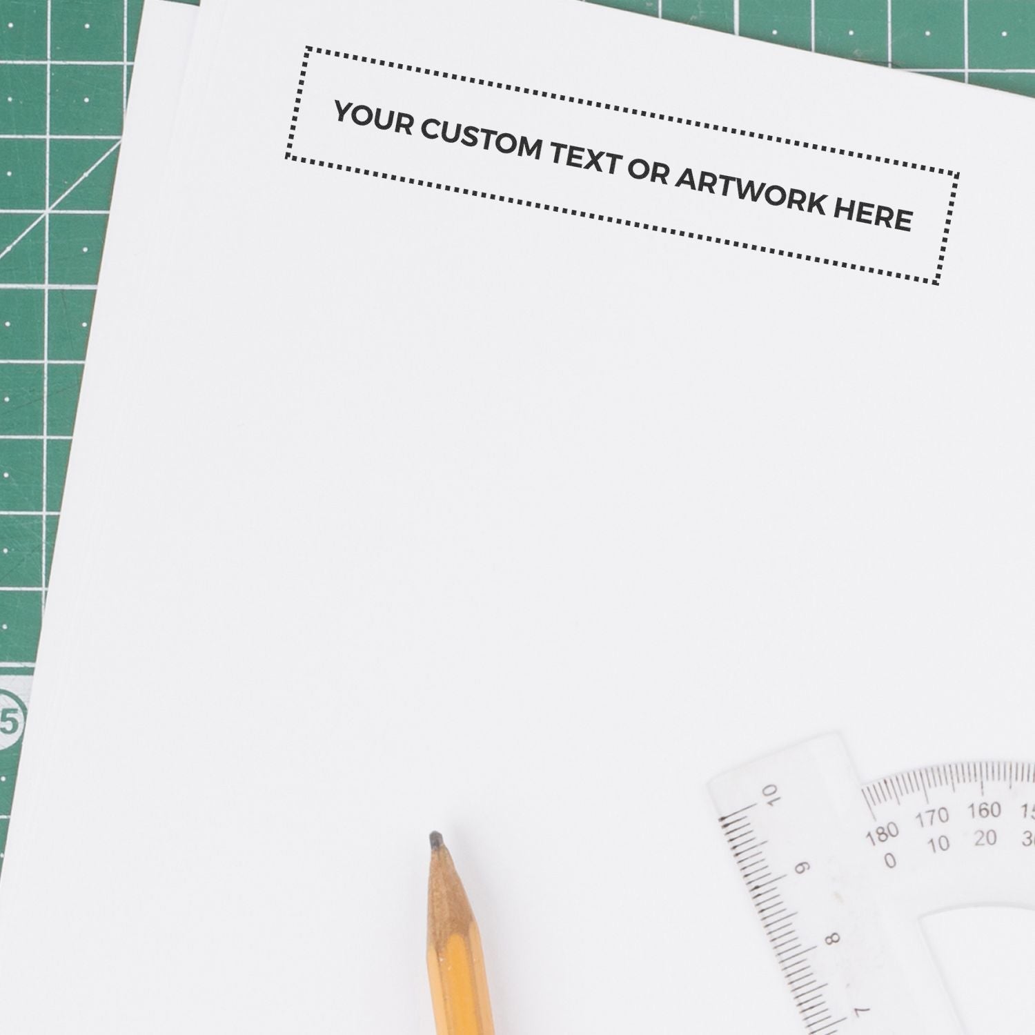 Custom Rubber Stamp Size 1 x 6 used on white paper with Your Custom Text or Artwork Here stamped, next to a pencil and ruler on a green cutting mat.
