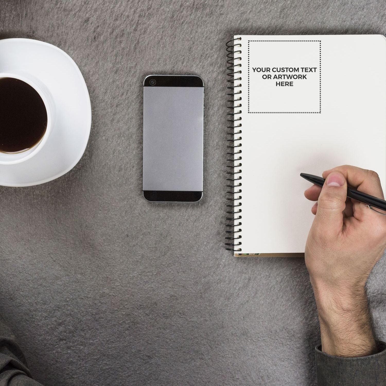 A hand writing in a notebook stamped with Your Custom Text or Artwork Here using a Custom Rubber Stamp Size 4 x 4, next to a phone and coffee.