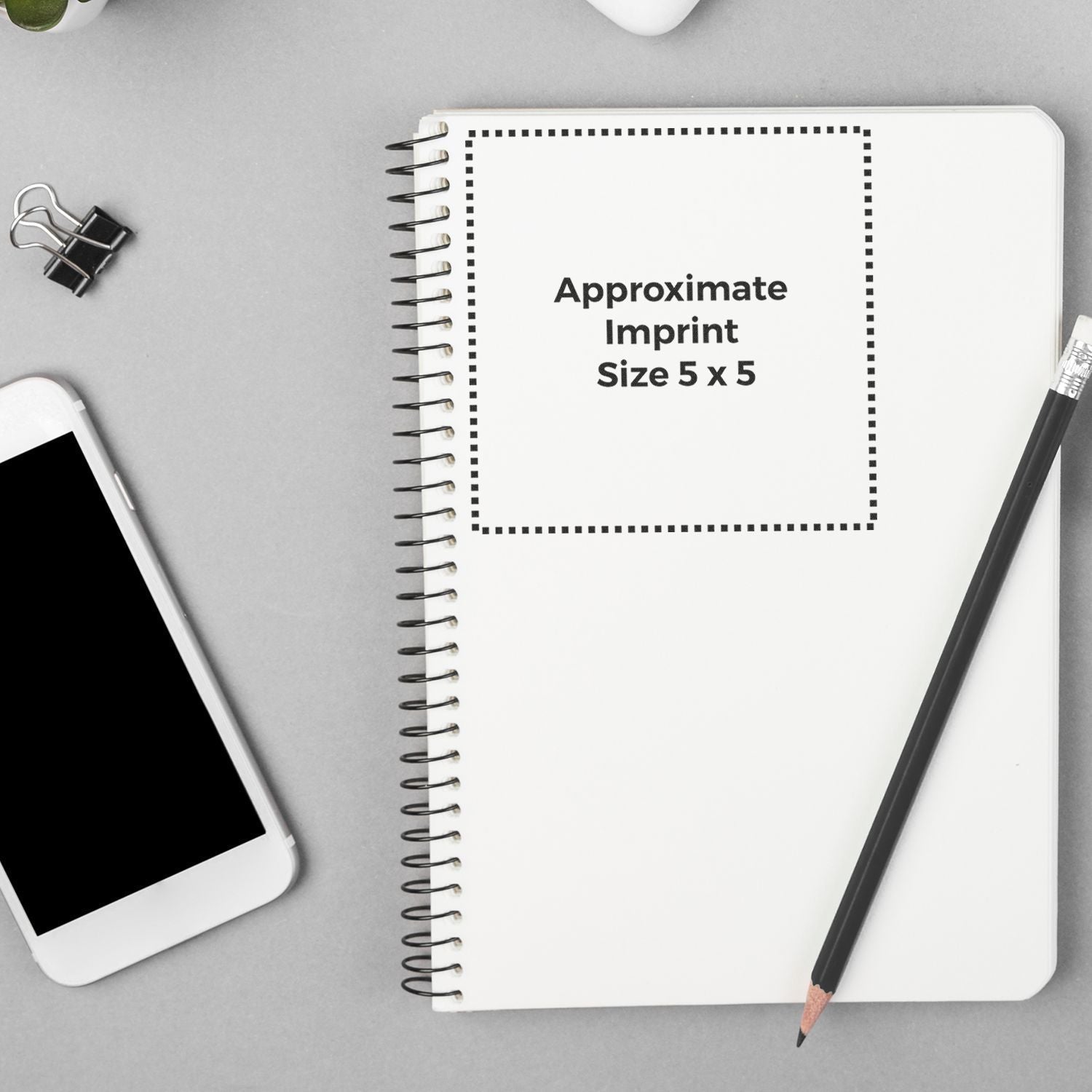 Custom Rubber Stamp Size 5 x 5 shown on a spiral notebook with a pencil, phone, and paper clip on a gray background.