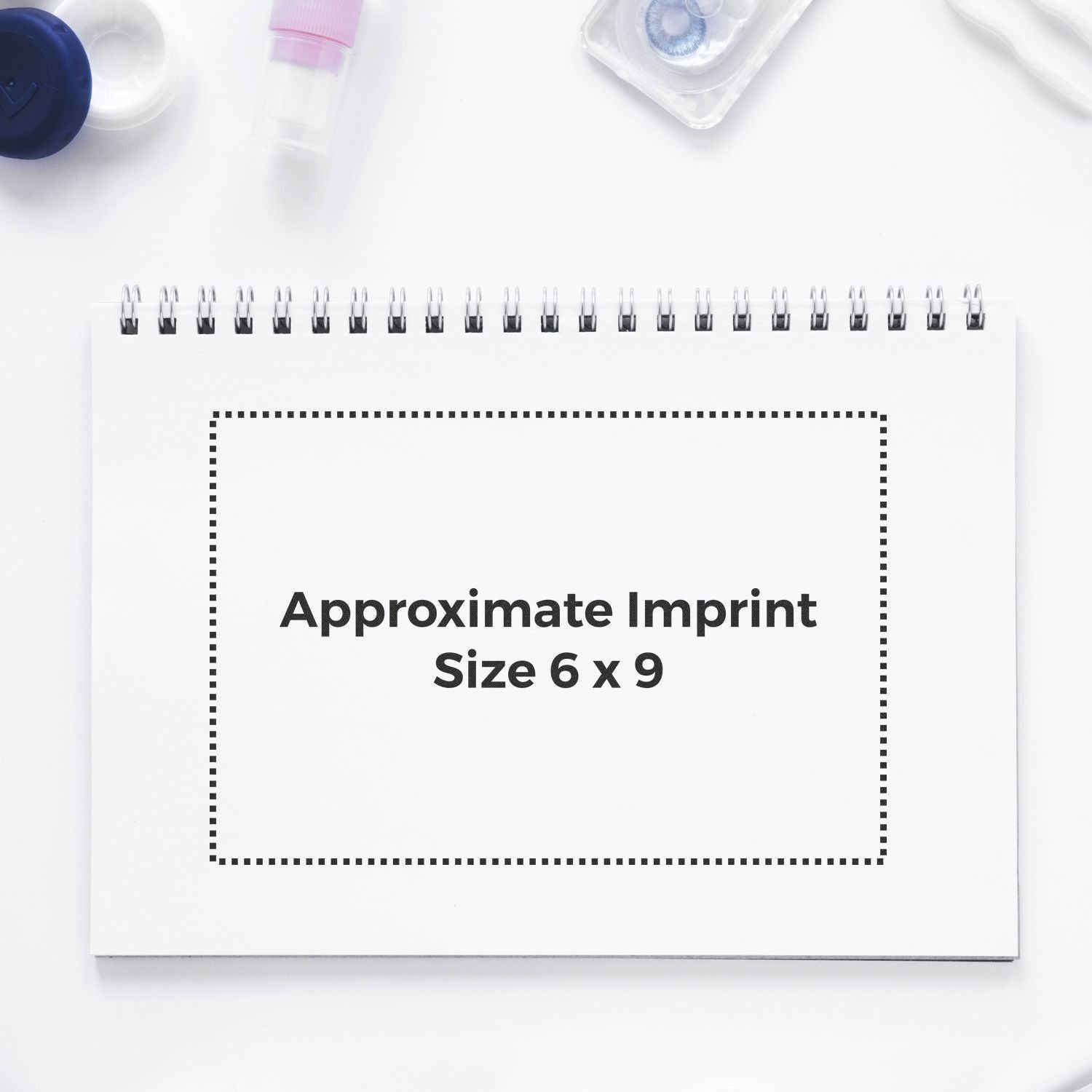 Custom Rubber Stamp Size 6 x 9 shown on a white spiral notebook with dotted outline indicating imprint area, surrounded by office supplies.