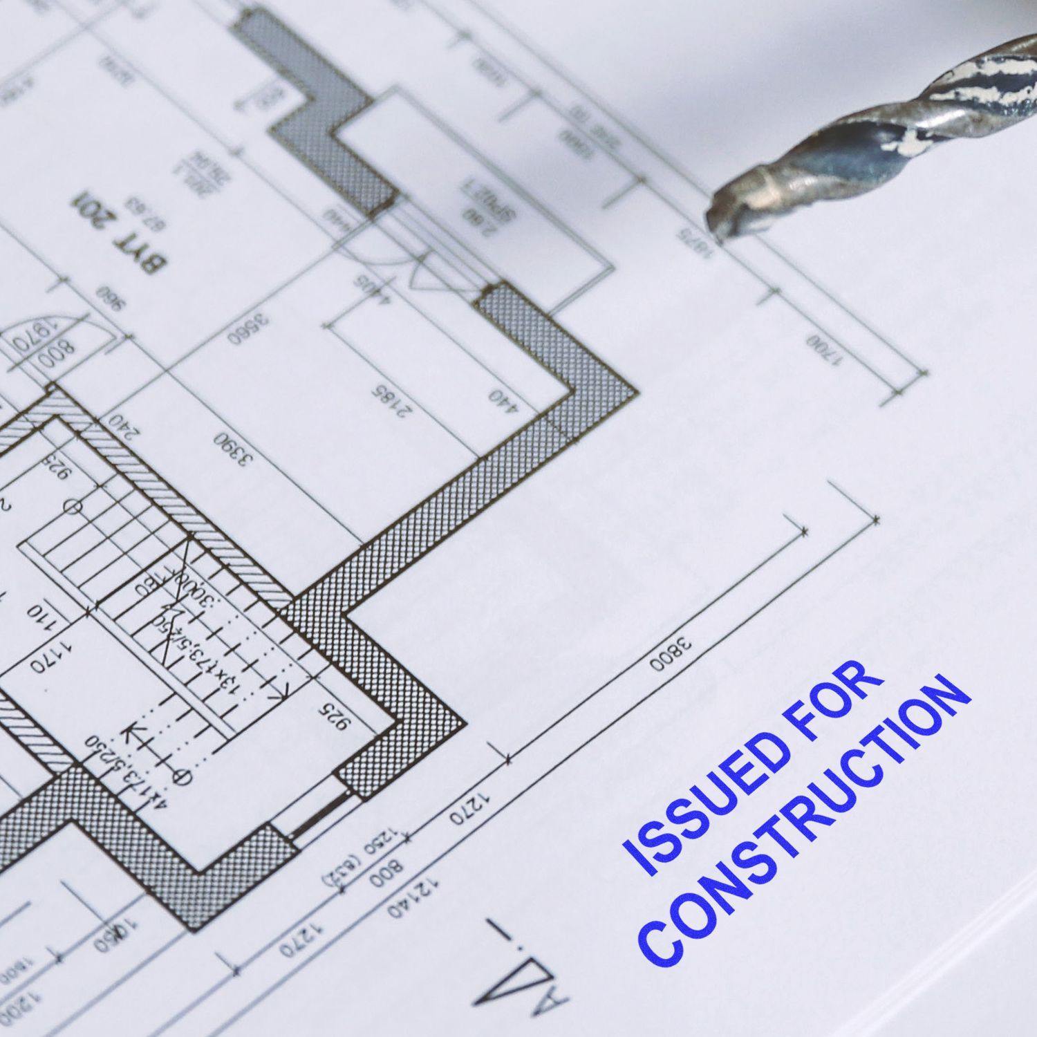 A blueprint with a Self Inking Issued for Construction Stamp marking in blue ink, next to a drill bit.