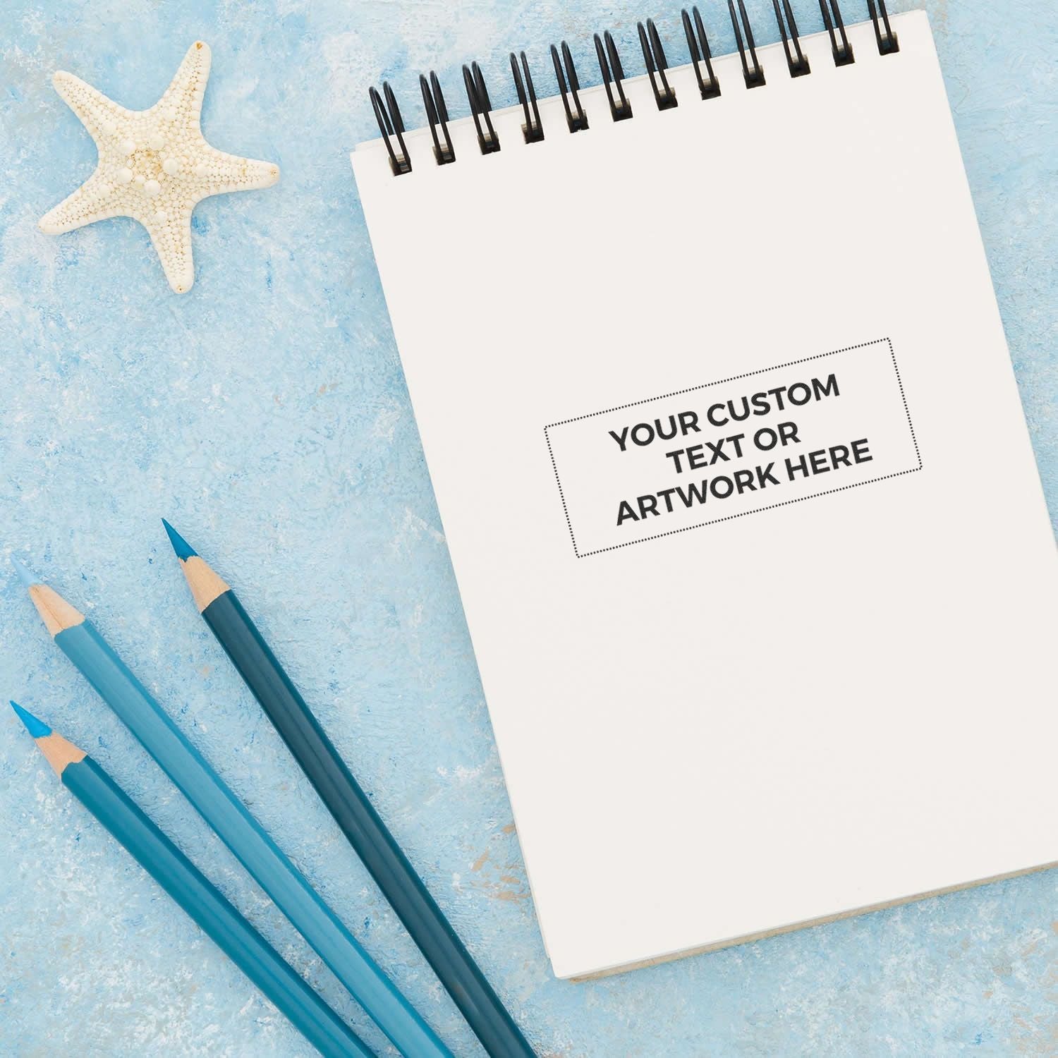 Slim Stamp 2773 Customized Pre-Inked Stamp 1-1/16 x 2-7/8 on a notepad with Your Custom Text or Artwork Here, next to pencils and a starfish.