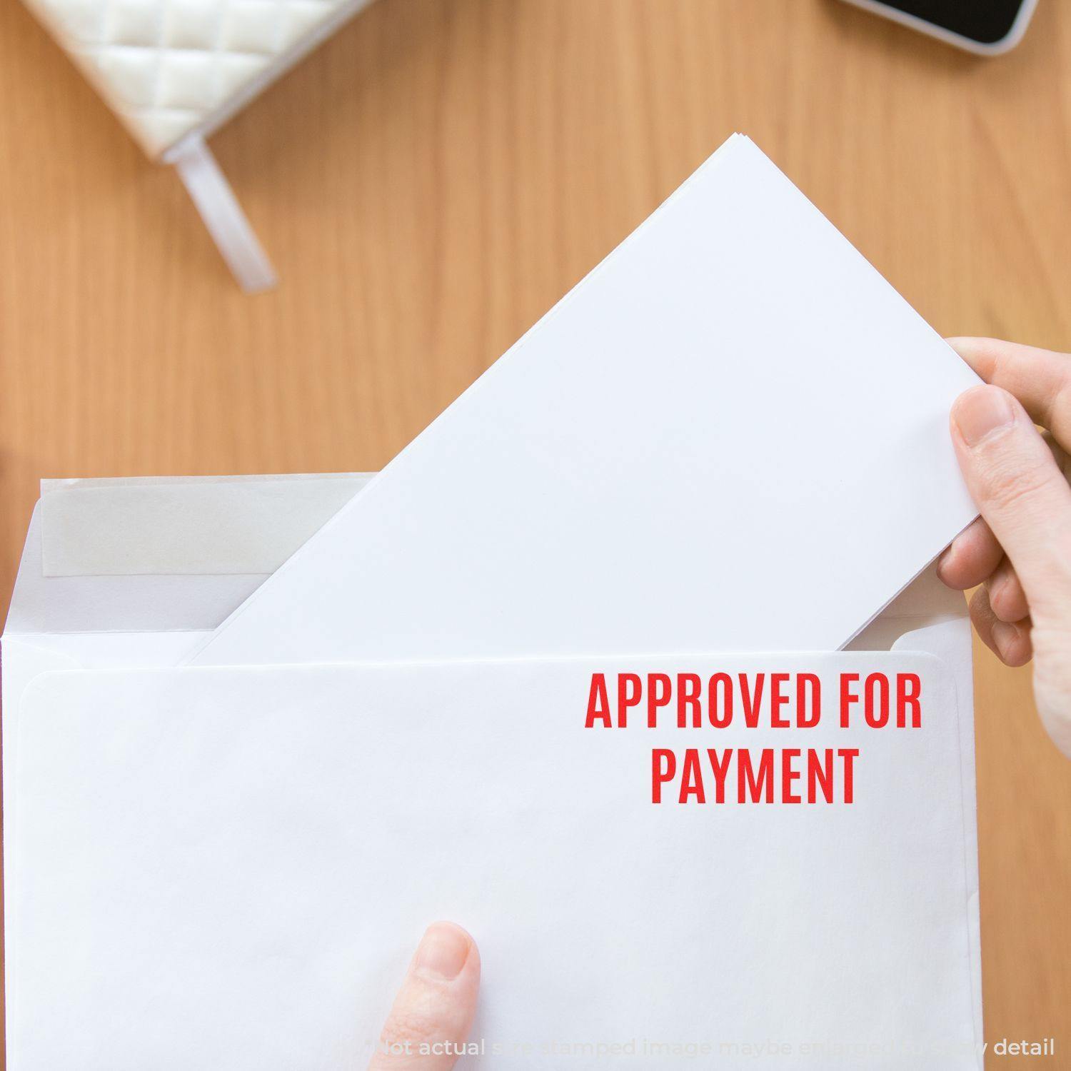 A hand placing an envelope into another envelope stamped with Approved for Payment using the Self Inking Additional Postage Required Stamp.