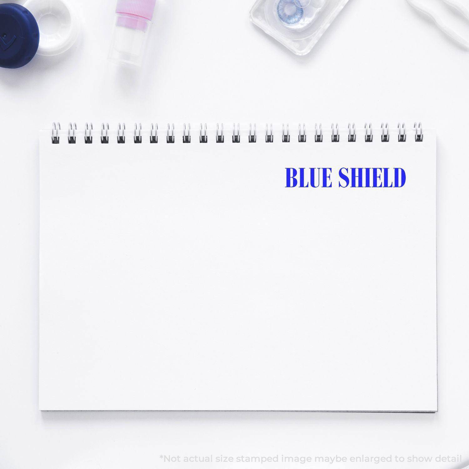 A Large Pre-Inked Blue Shield Stamp imprint on a white notepad, surrounded by office supplies.