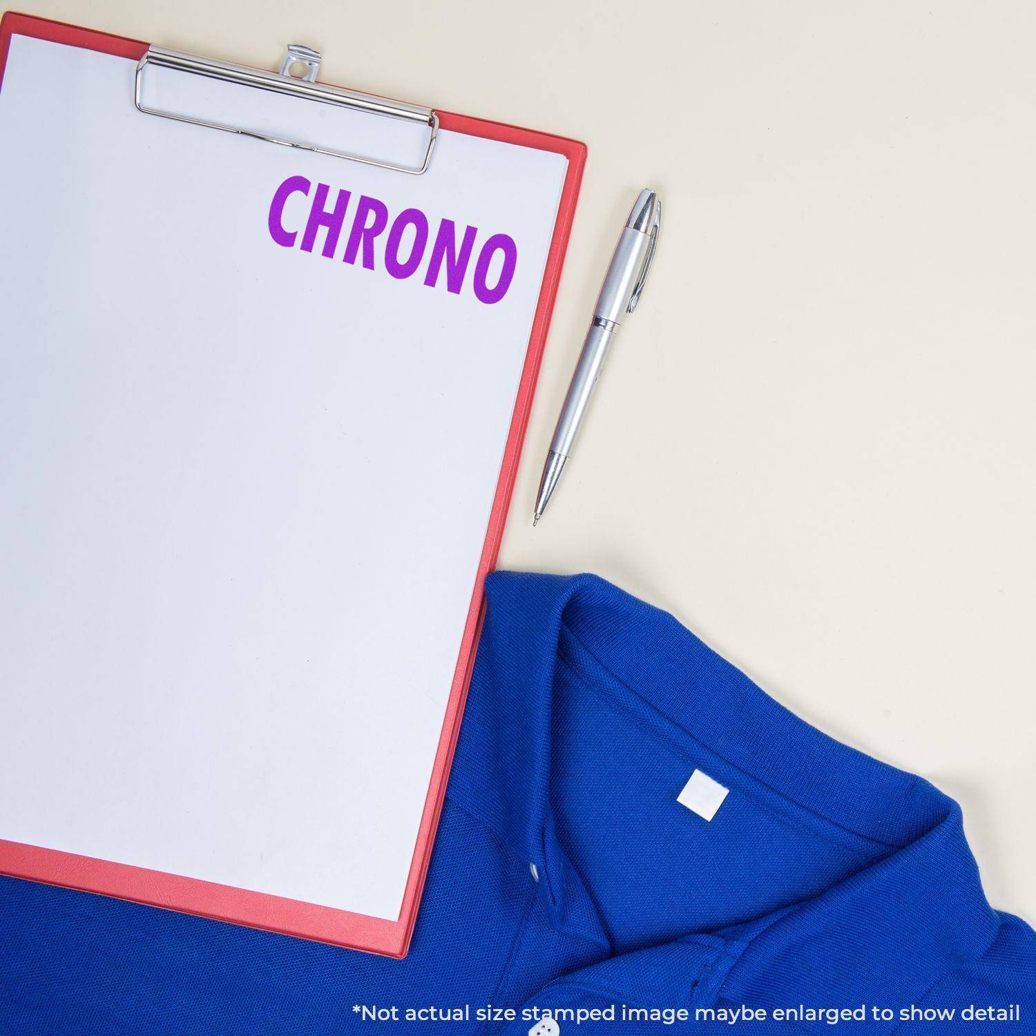 Clipboard with CHRONO stamped in purple using the Large Self Inking Chrono Stamp, next to a pen and blue shirt on a light surface.