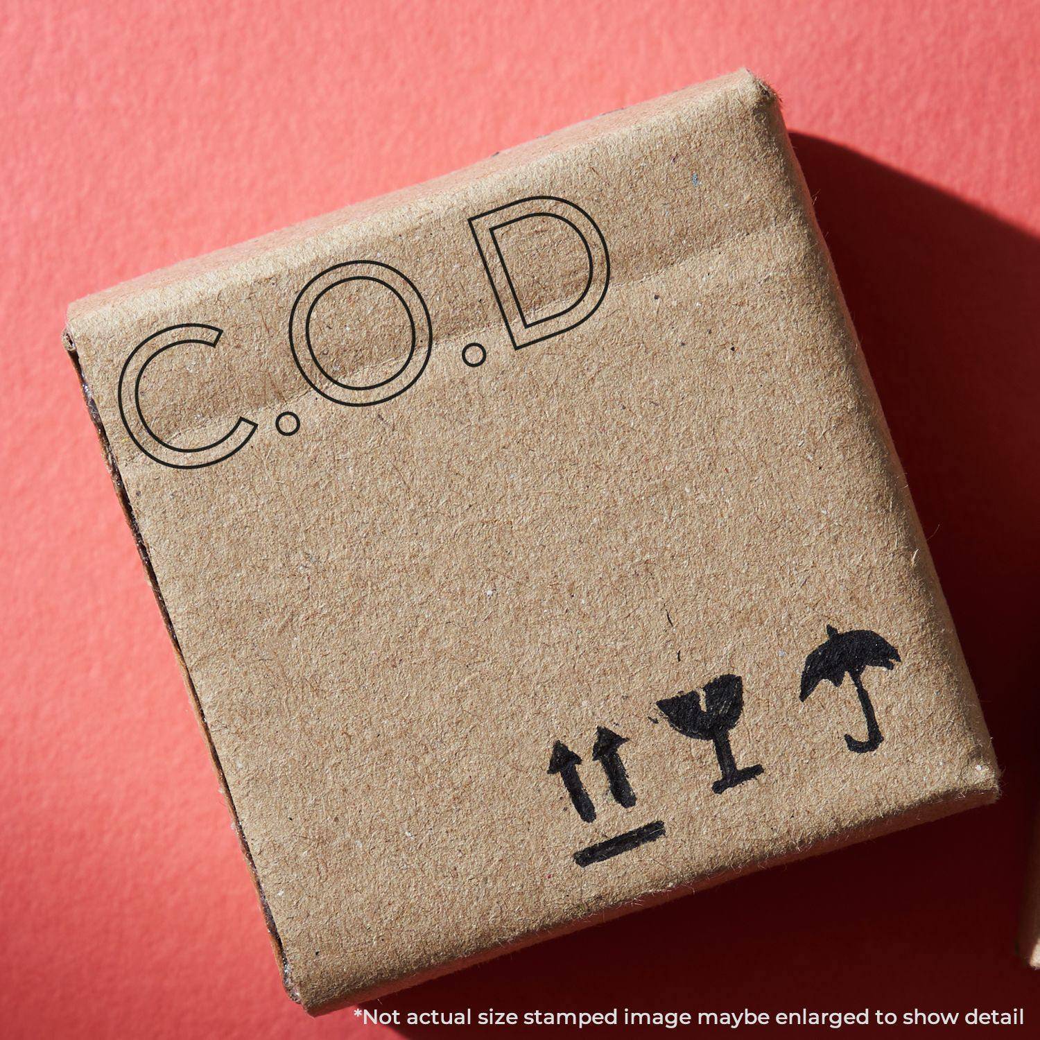 A cardboard box stamped with C.O.D using the Large Self Inking COD Outline Stamp, placed on a red surface. The box also features handling symbols for fragile, this side up, and keep dry.