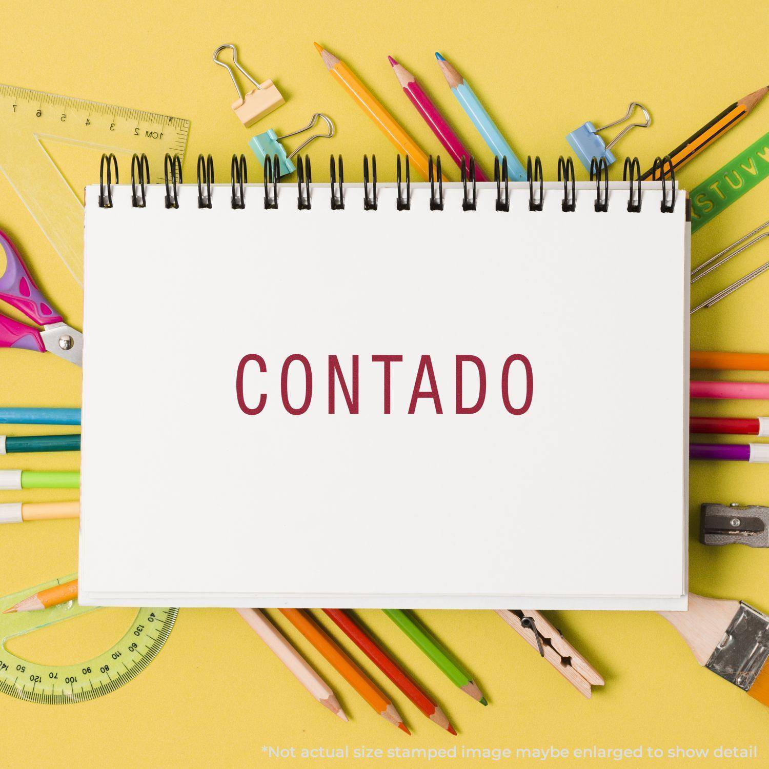 A Large Self Inking Contado Stamp is shown in use, stamping the word CONTADO on a white notebook page. The notebook is surrounded by colorful stationery items on a yellow background.